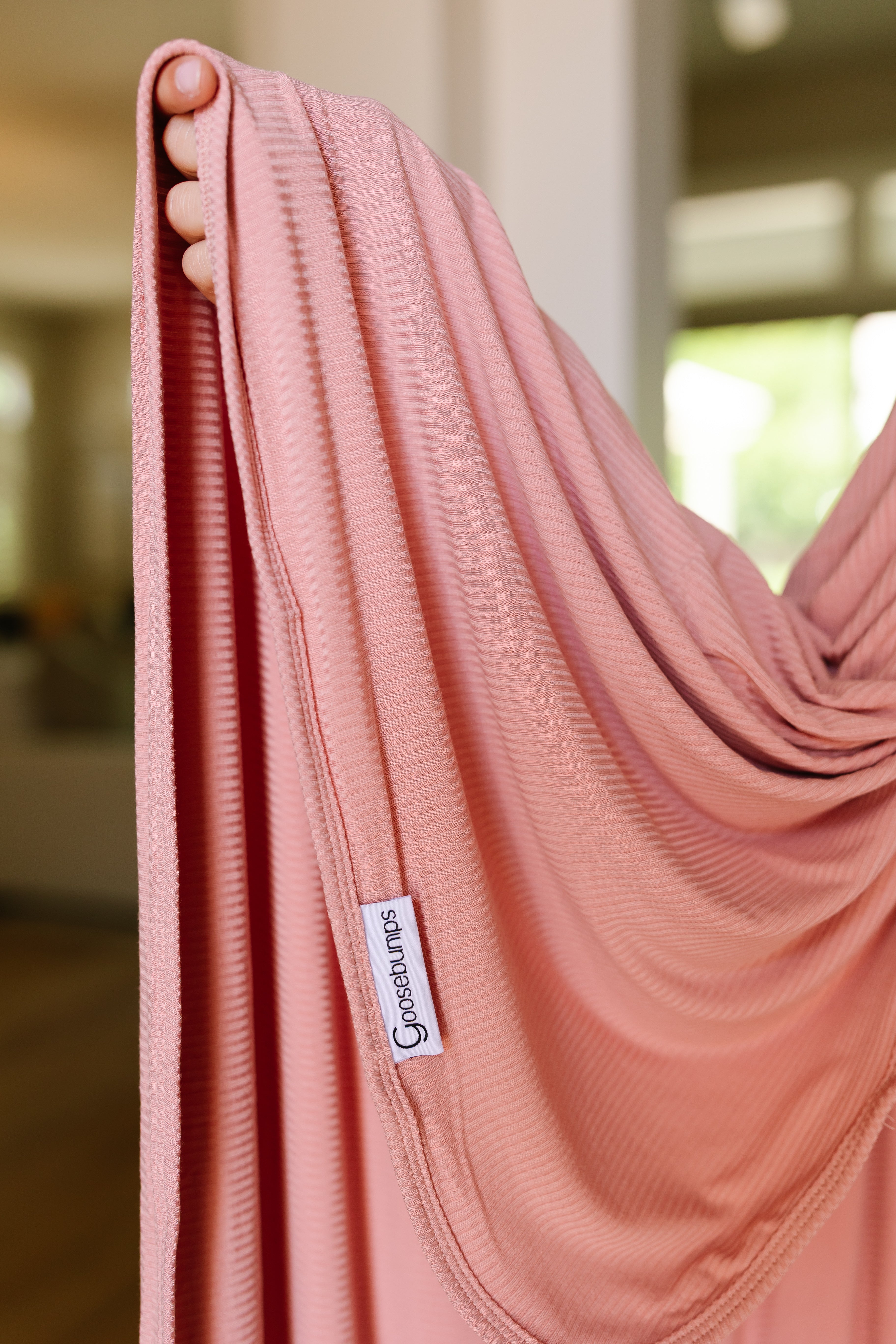 A hand holds up the peony, a soft ribbed pink blanket with a white Gossie Lups label. The background shows an indoor space with blurred windows and furniture.