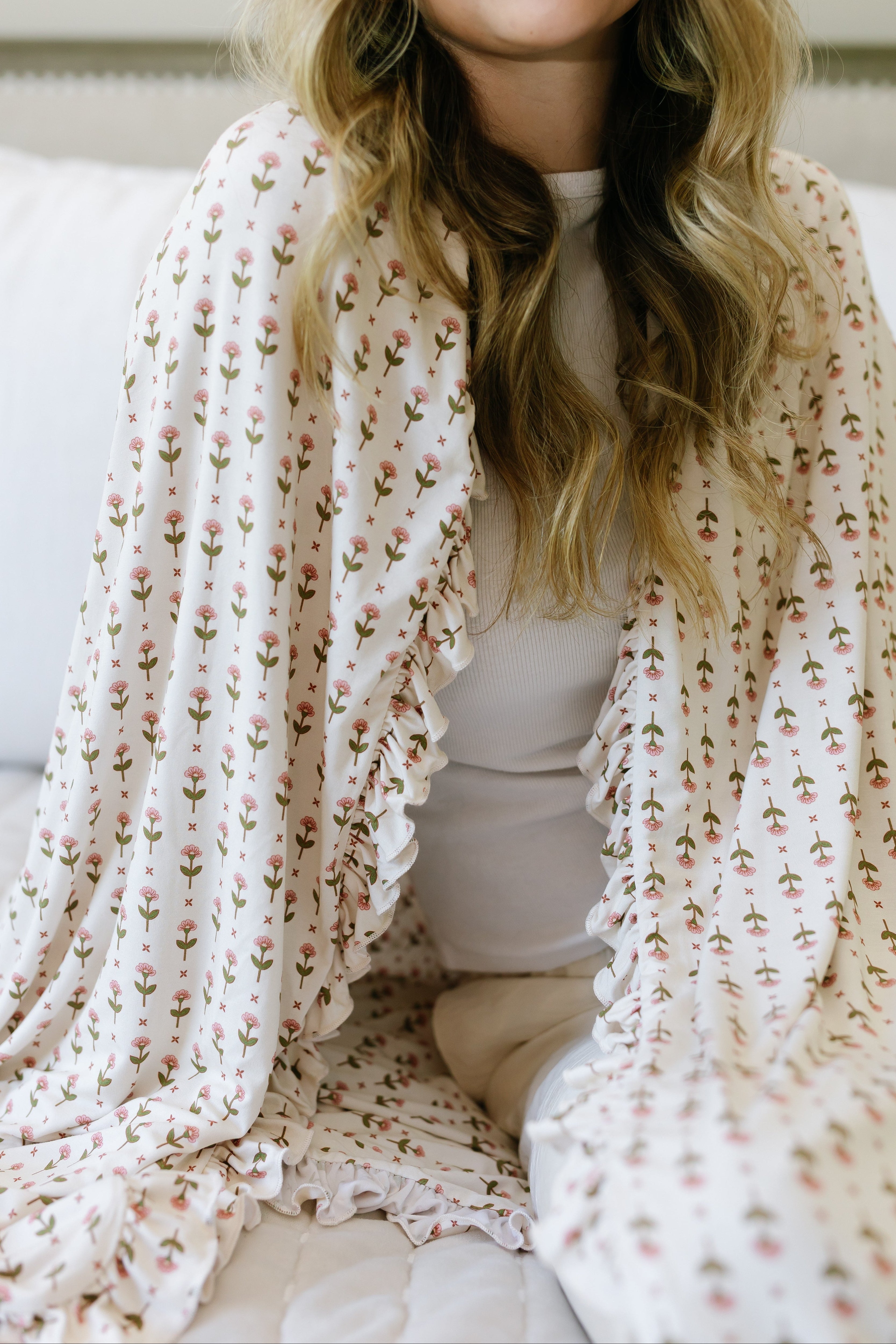 A woman with wavy blonde hair sits on a white bed, wrapped in the tulip / ruffle blanket with its delicate floral pattern over a white outfit. Her face is partly out of frame, adding to the scene's soft, cozy ambiance.