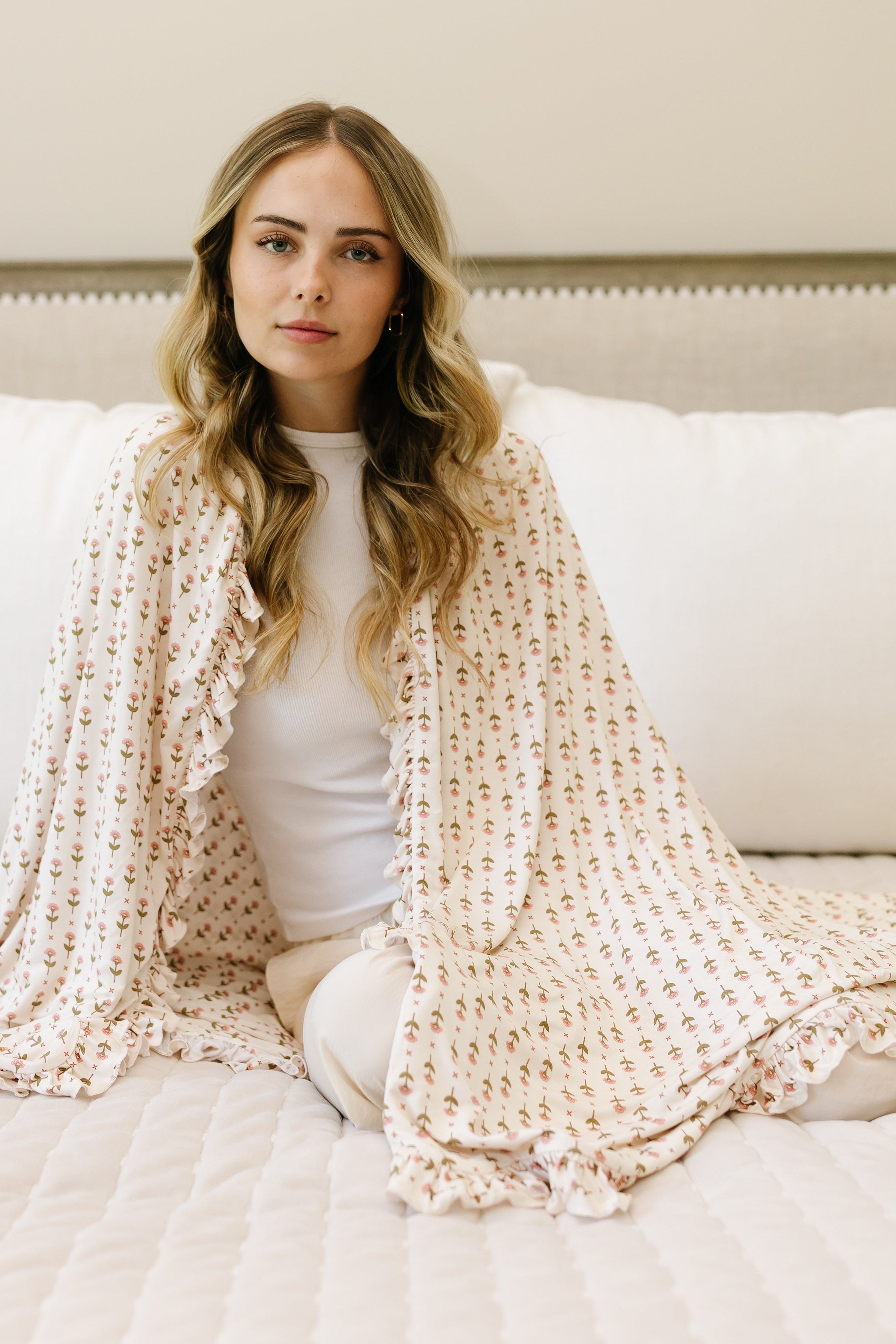 A woman with long, wavy blonde hair sits cross-legged on a bed, wrapped in the tulip / ruffle patterned shawl and luxurious blanket, with white pillows and a neutral-toned headboard in the background.