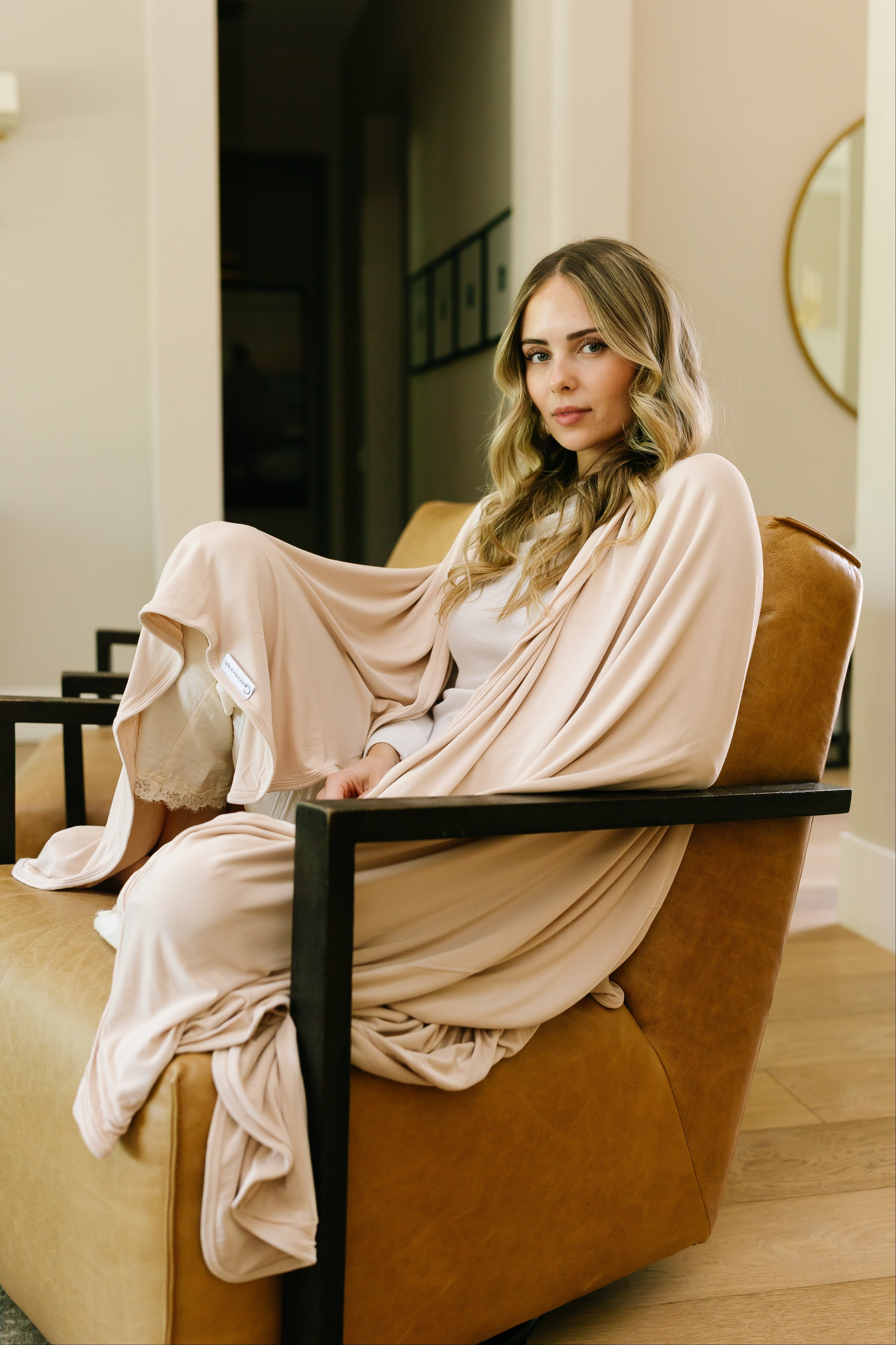 A woman with long, wavy blonde hair wearing a light beige robe sits on a tan leather armchair, wrapped in the luxurious vanilla cream blanket, in a modern, softly lit room with neutral decor.