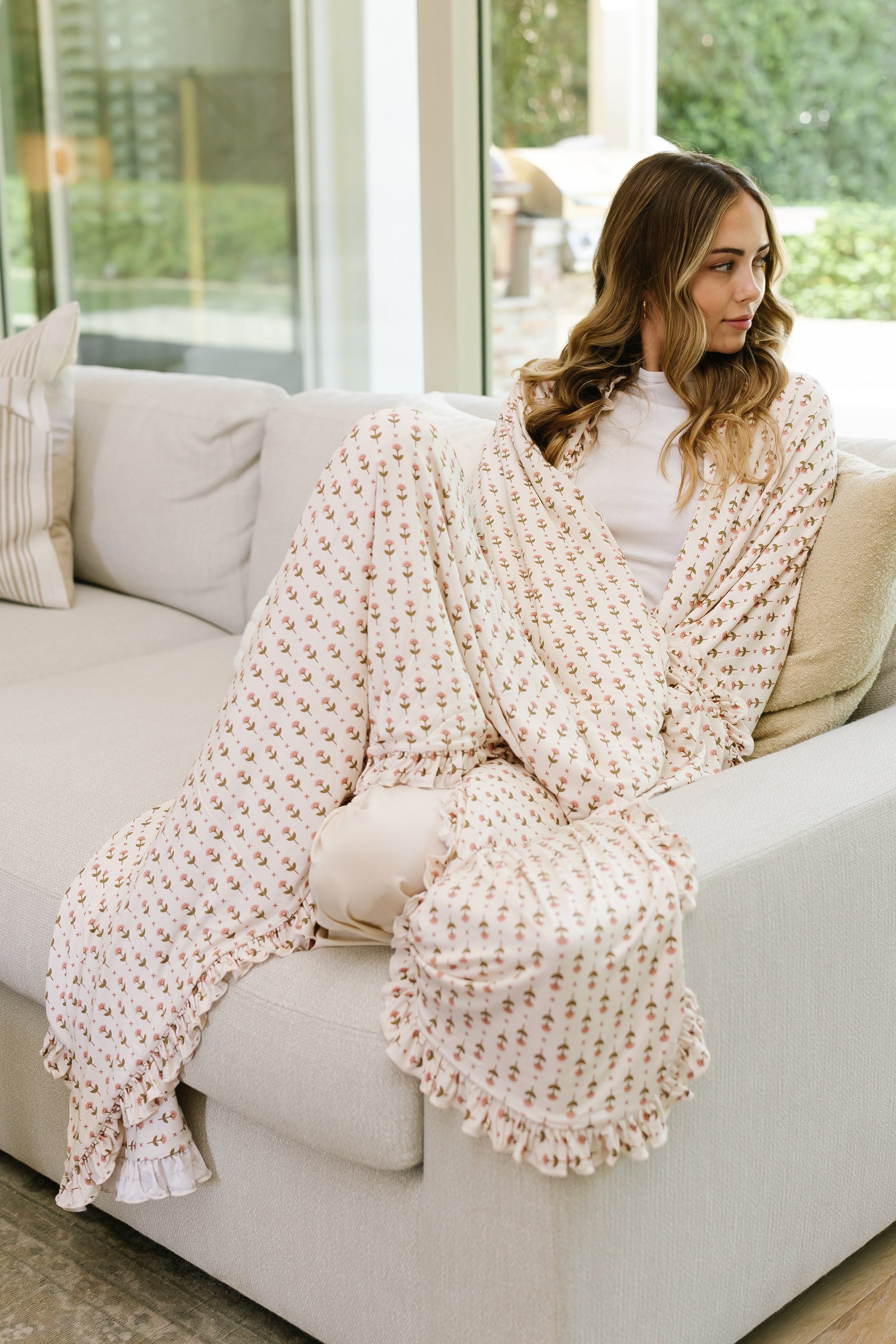 A woman with long hair sits on a light couch, wrapped in the tulip / ruffle blanket featuring a small floral pattern. She wears a white top and light pants, looking to the side as greenery is visible through large windows behind her.