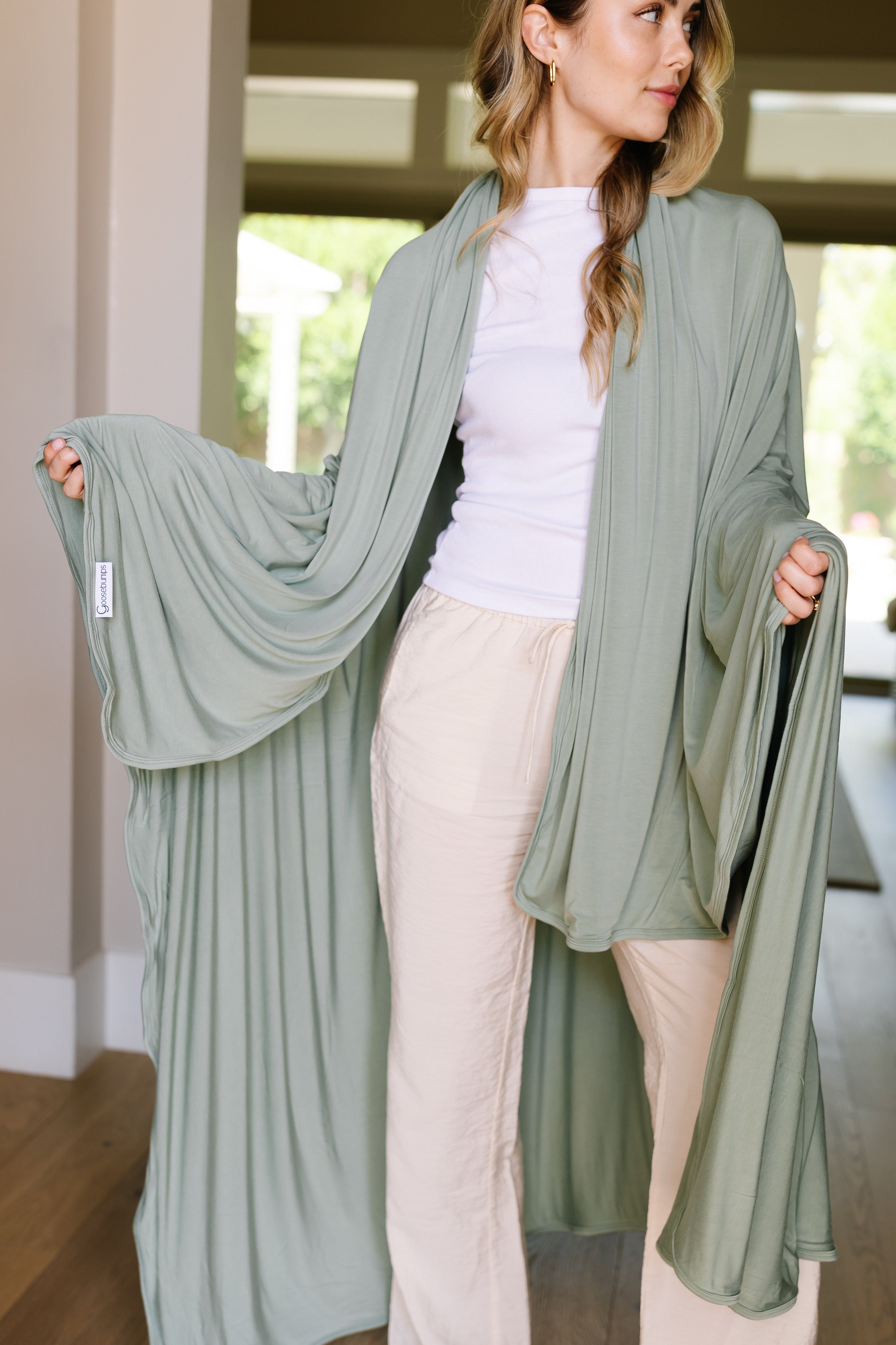 A woman with long blonde hair smiles softly as she stands indoors, modeling "basil," a light green, flowing robe with wide sleeves over a white top and beige pants. She holds out the sleeves, showcasing the modern blanket-inspired design.