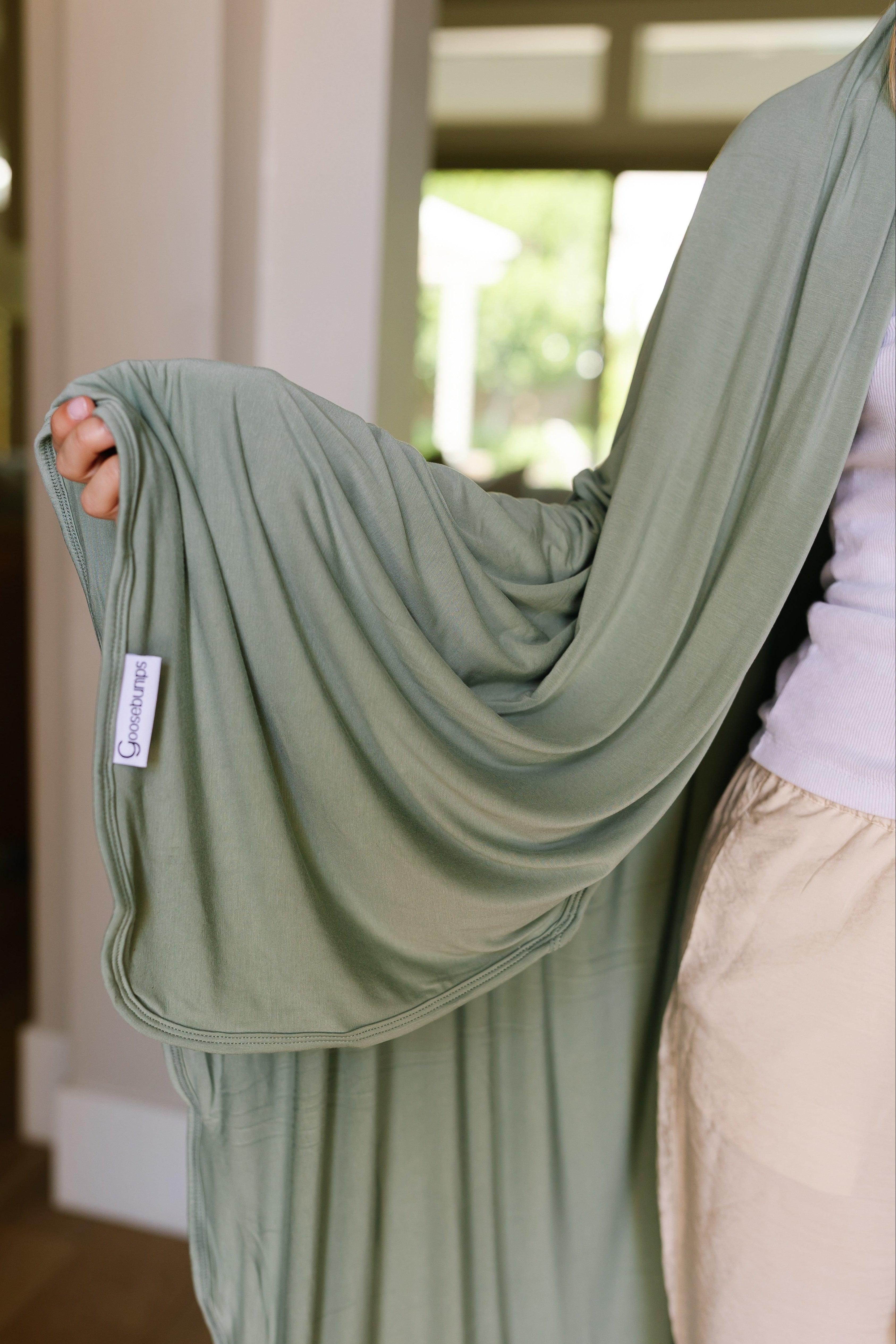 A person holds up a luxurious basil blanket with a sewn-in Saranoni label. They wear a white top and beige pants, while bright windows light up the modern background.