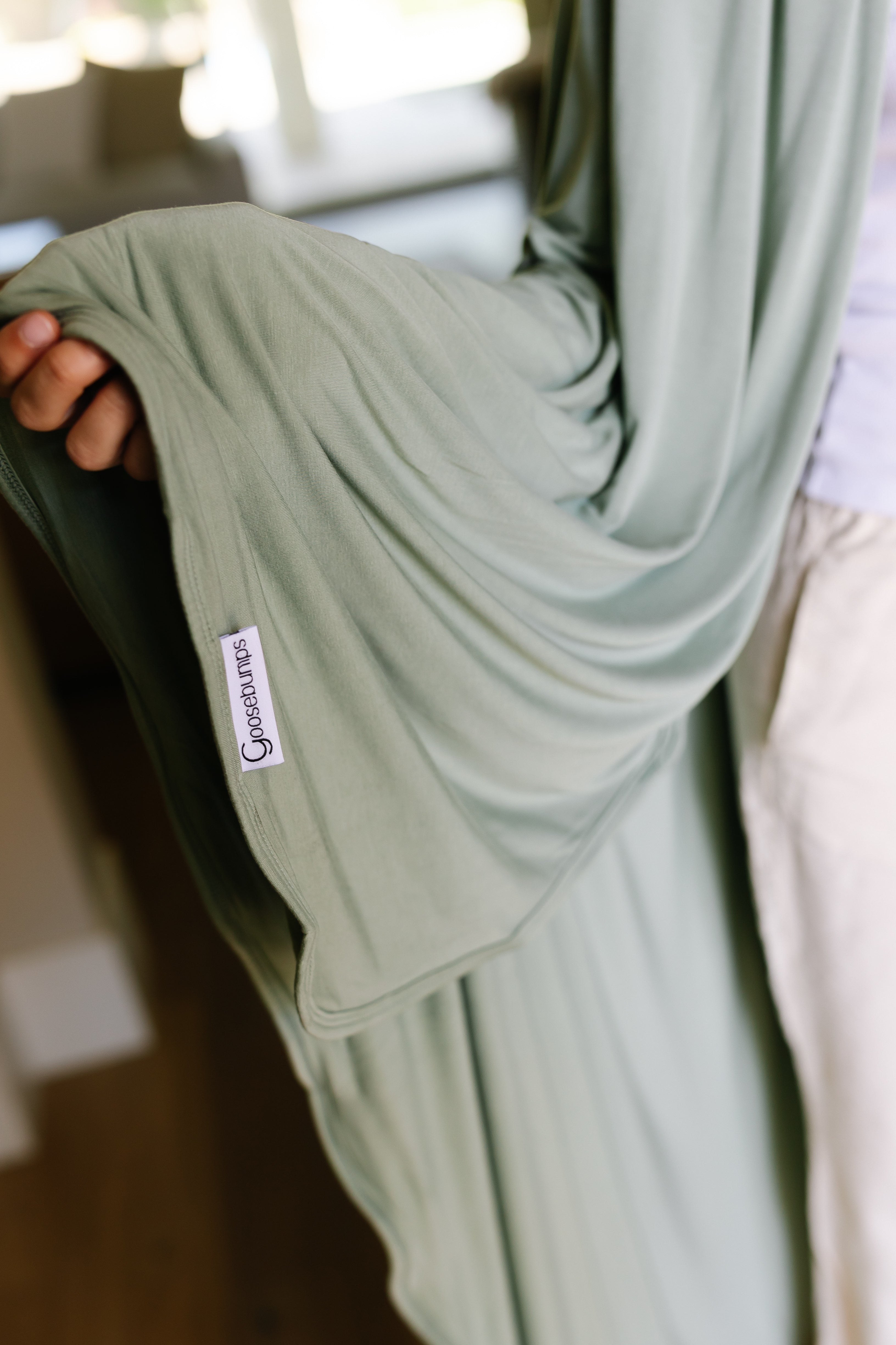 A person holds out the loose, flowing sleeve of a light green "basil" robe with a sewn-in CaspBaby tag, standing indoors on a wooden floor—the soft fabric as gentle as a bamboo blanket.