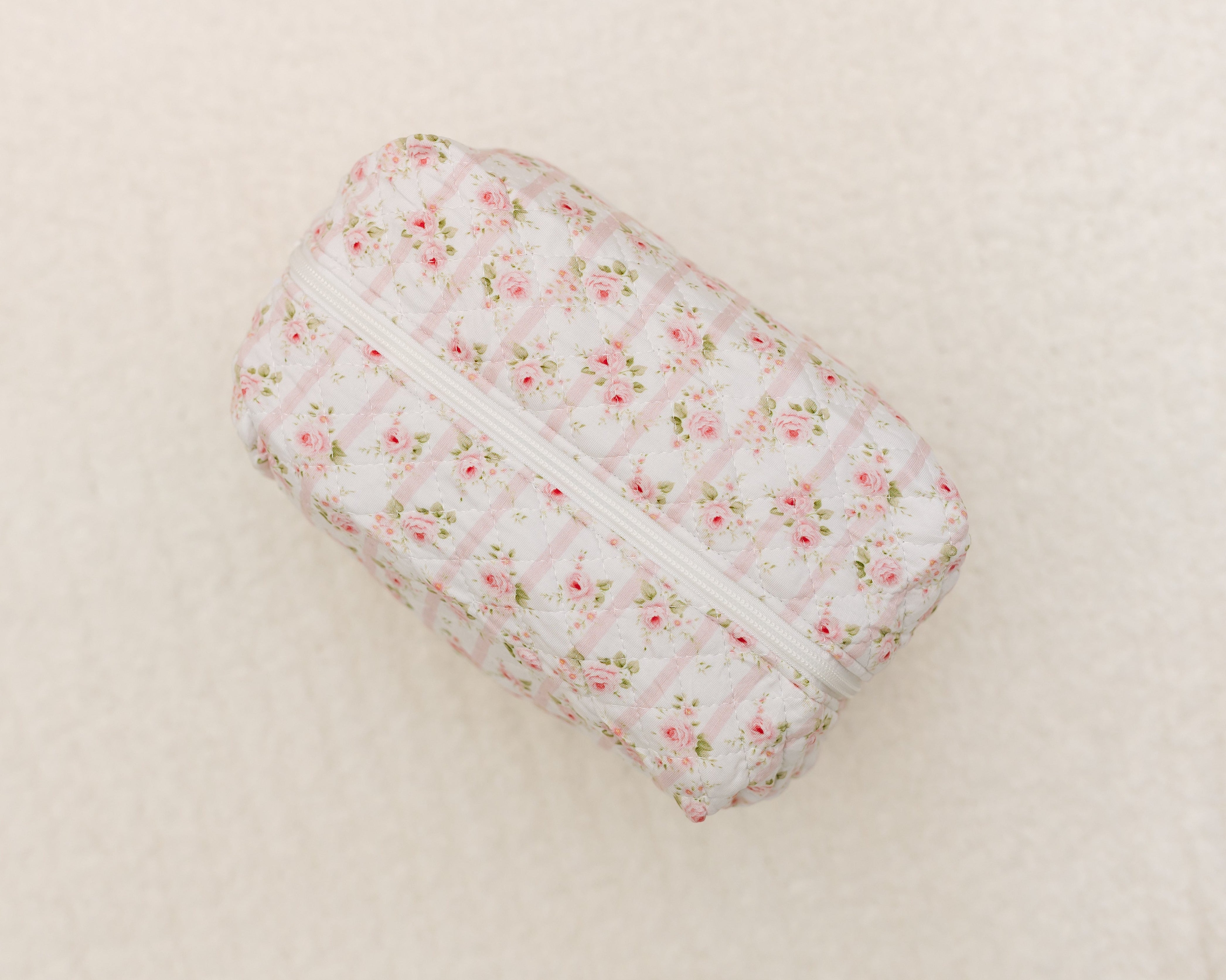 The rosie / travel bag is a rectangular quilted pouch with a zipper, showcasing a pink floral print on white and displayed on a light beige textured background.