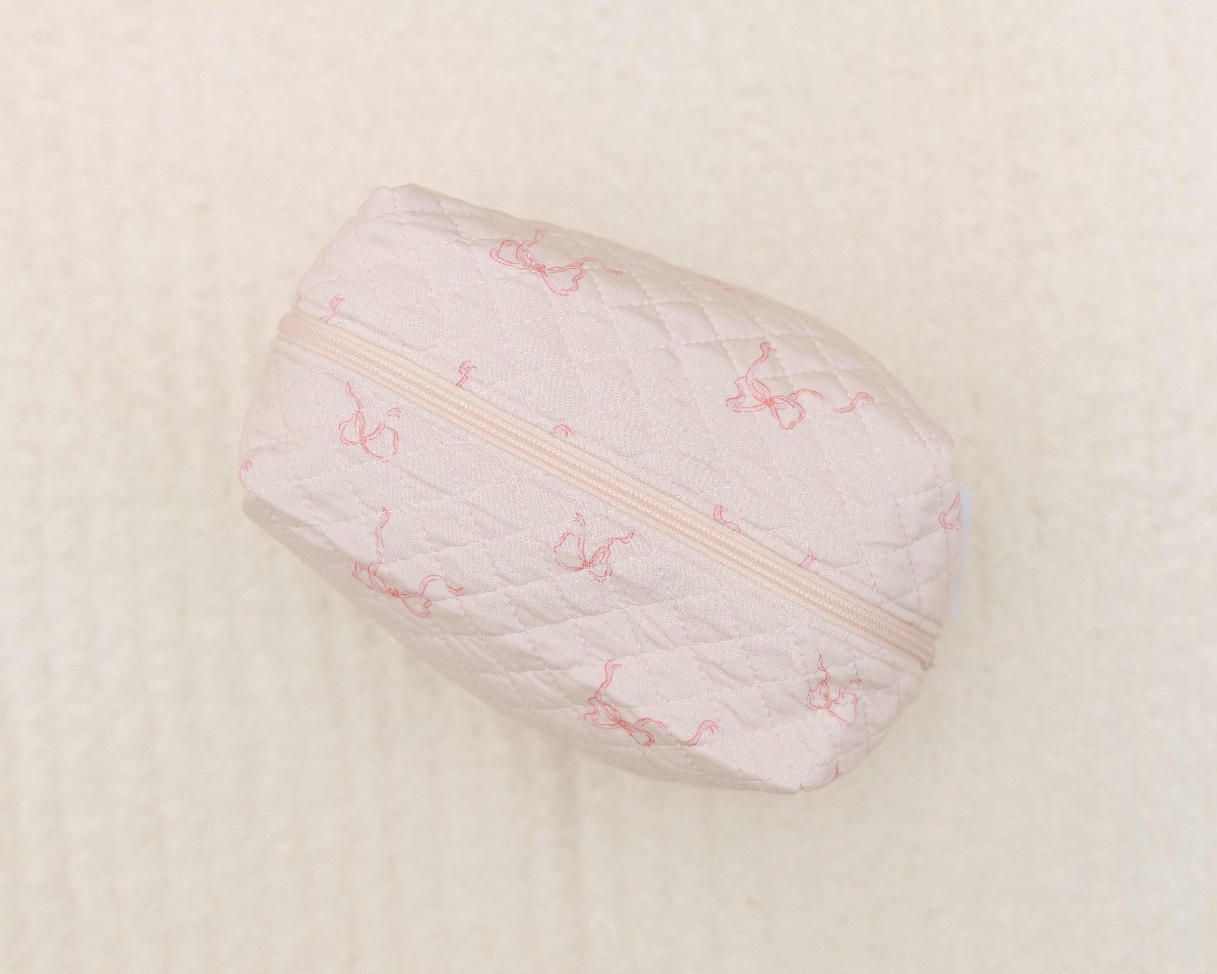The ballerina / travel bag is a small, quilted, light pink travel accessory with a subtle cherry and bow pattern, ideal for carrying essentials. It’s shown on a textured cream-colored surface.