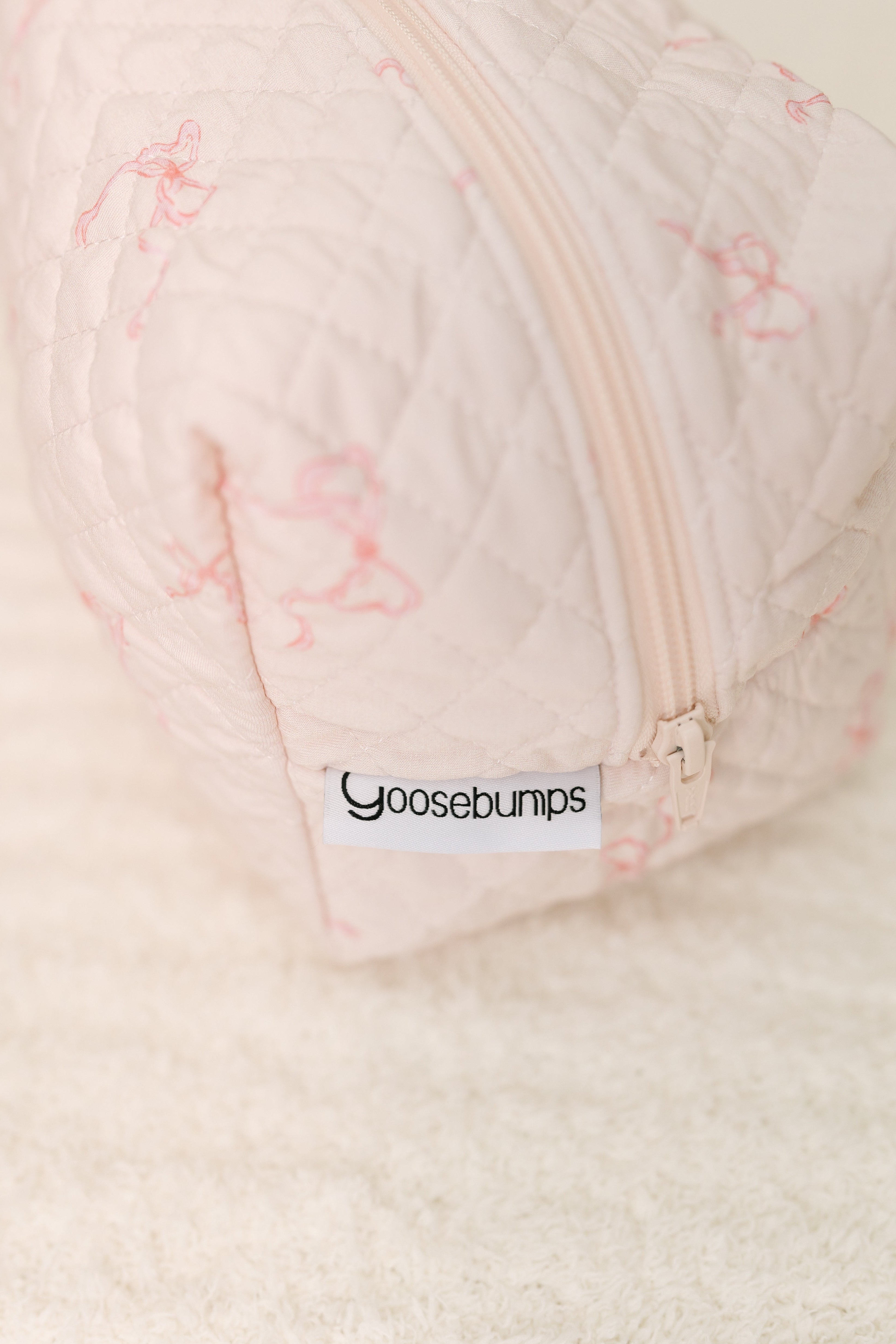 A close-up of the quilted, light pink ballerina / travel bag with a white goosebumps label. This chic pouch for travel essentials rests on a soft, textured white surface.