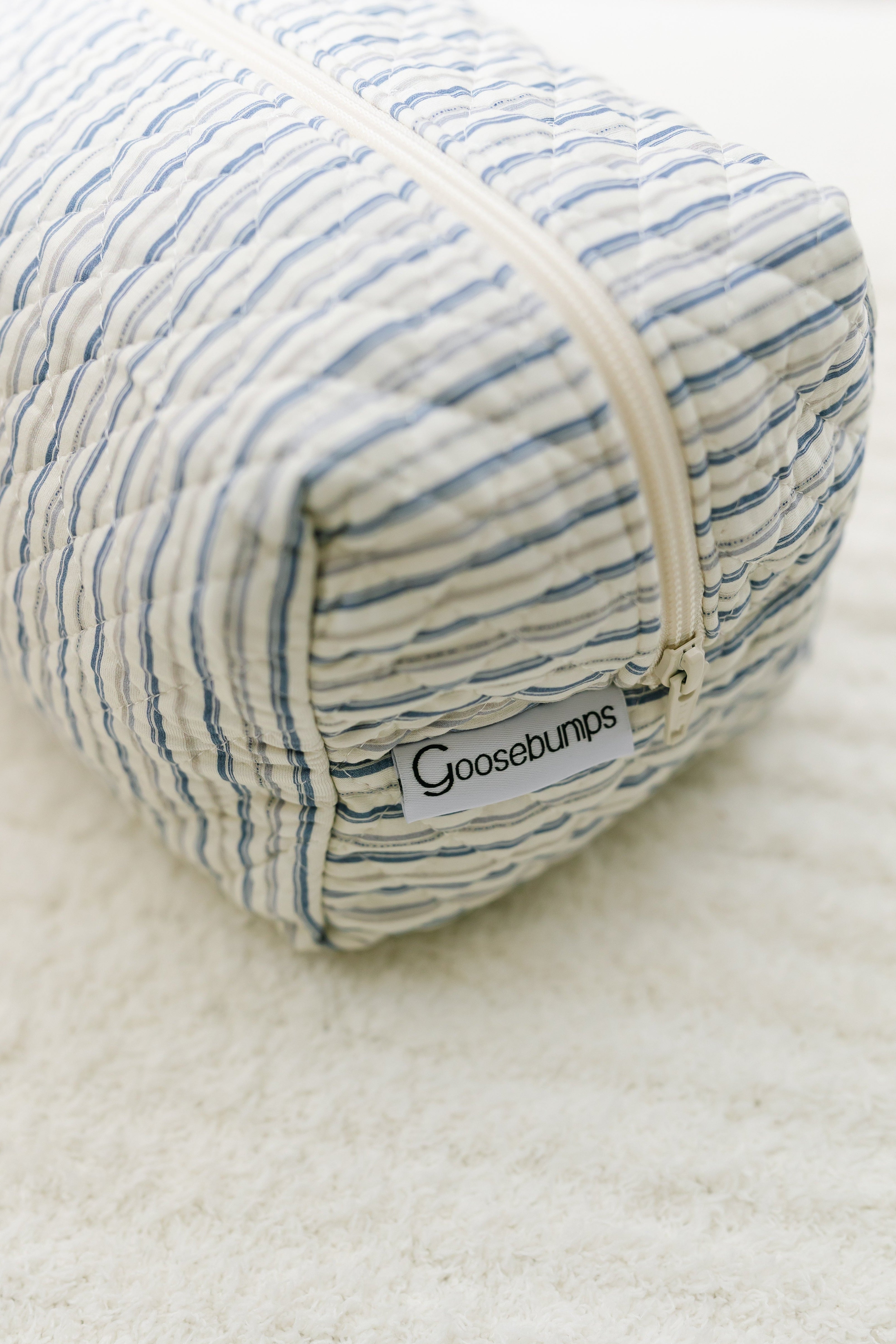 The beach house / travel bag is a quilted Goosebumps Travel Bag with blue and white wavy stripes, a zipper, and a Goosebumps label—stylish and perfect for your travel essentials.