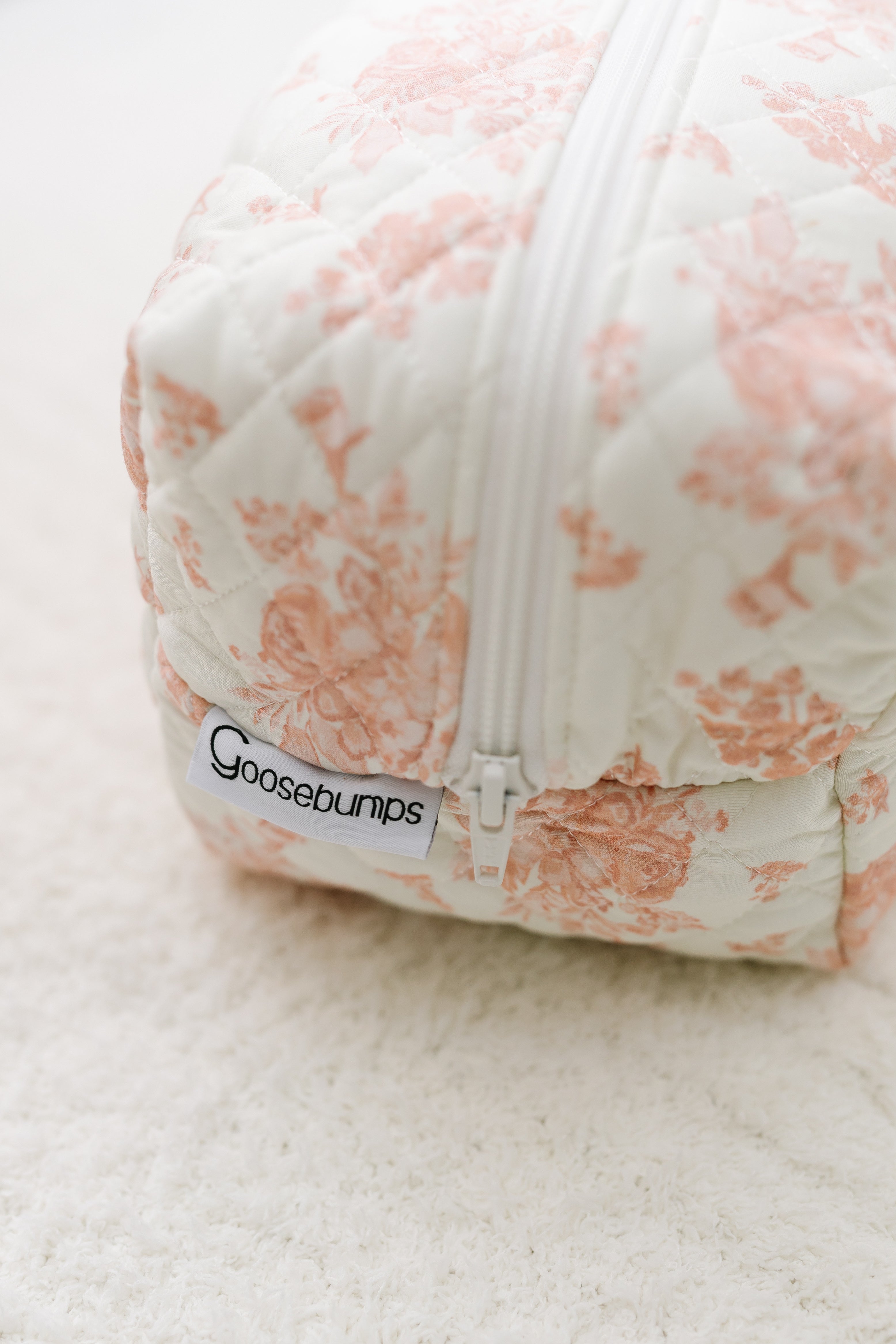 A close-up of the romance / travel bag—a quilted fabric bag with a white zipper, pink floral print, and label—resting on a light, textured surface.