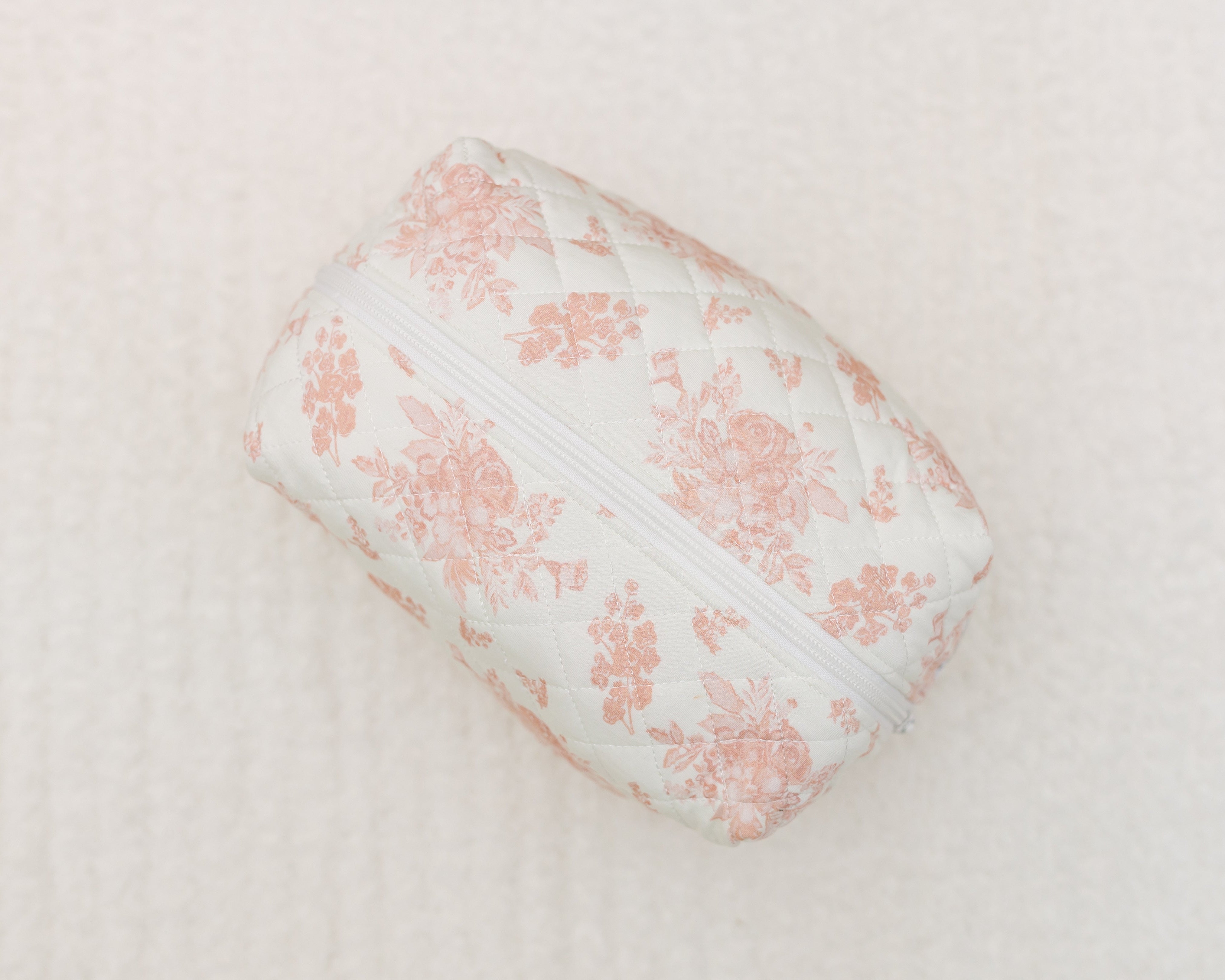 The romance / travel bag is a small quilted pouch with a pink floral print and white zipper, shown on a soft light surface—ideal as a cute travel bag for your essentials.