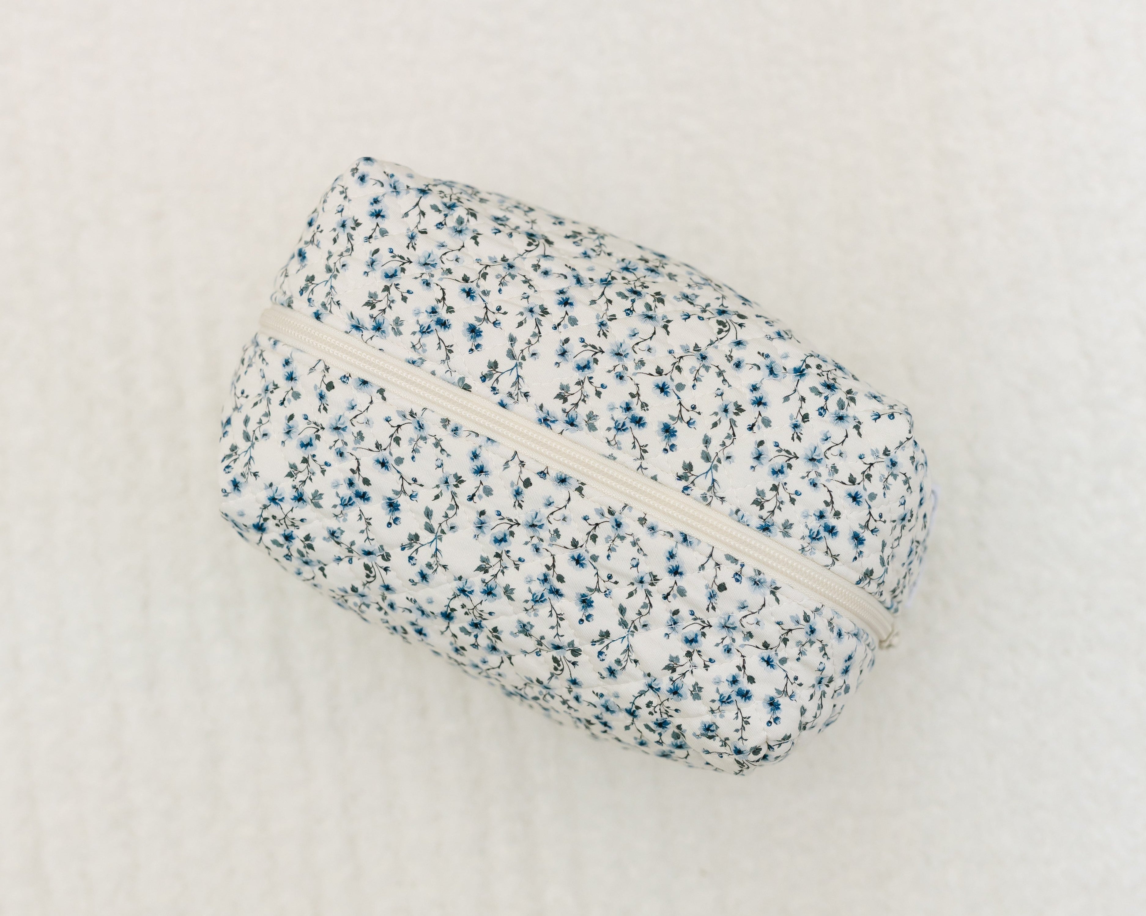 The cottage / travel bag is a rectangular pouch with a white background, featuring blue and black floral prints and a white zipper, displayed on a textured white surface—ideal for stylish, organized travel.