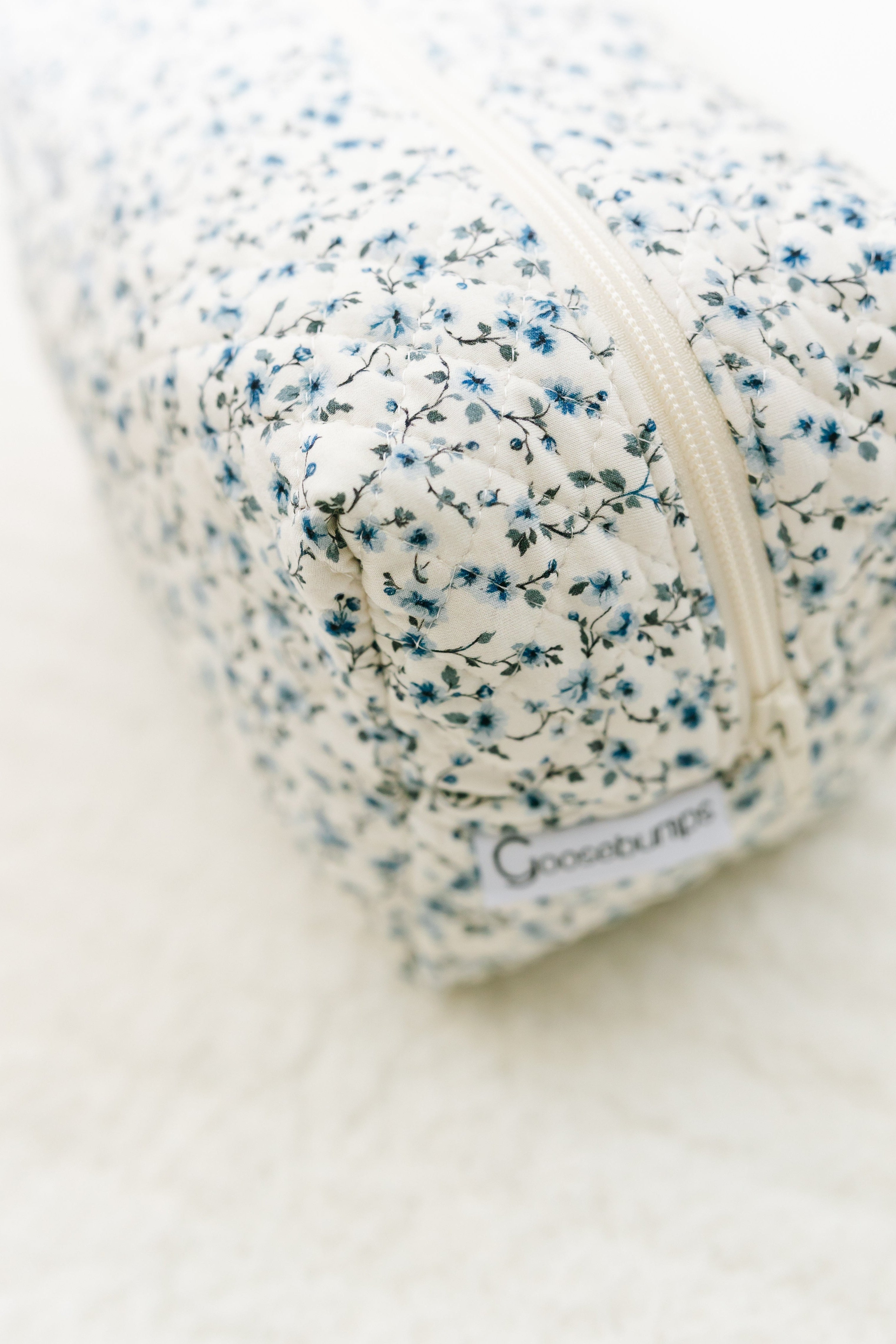 A rectangular cottage / travel bag with a blue and white floral print and a cream zipper rests on a soft white surface. A Goosebumps label at one end highlights this as the ideal cottage / travel bag.