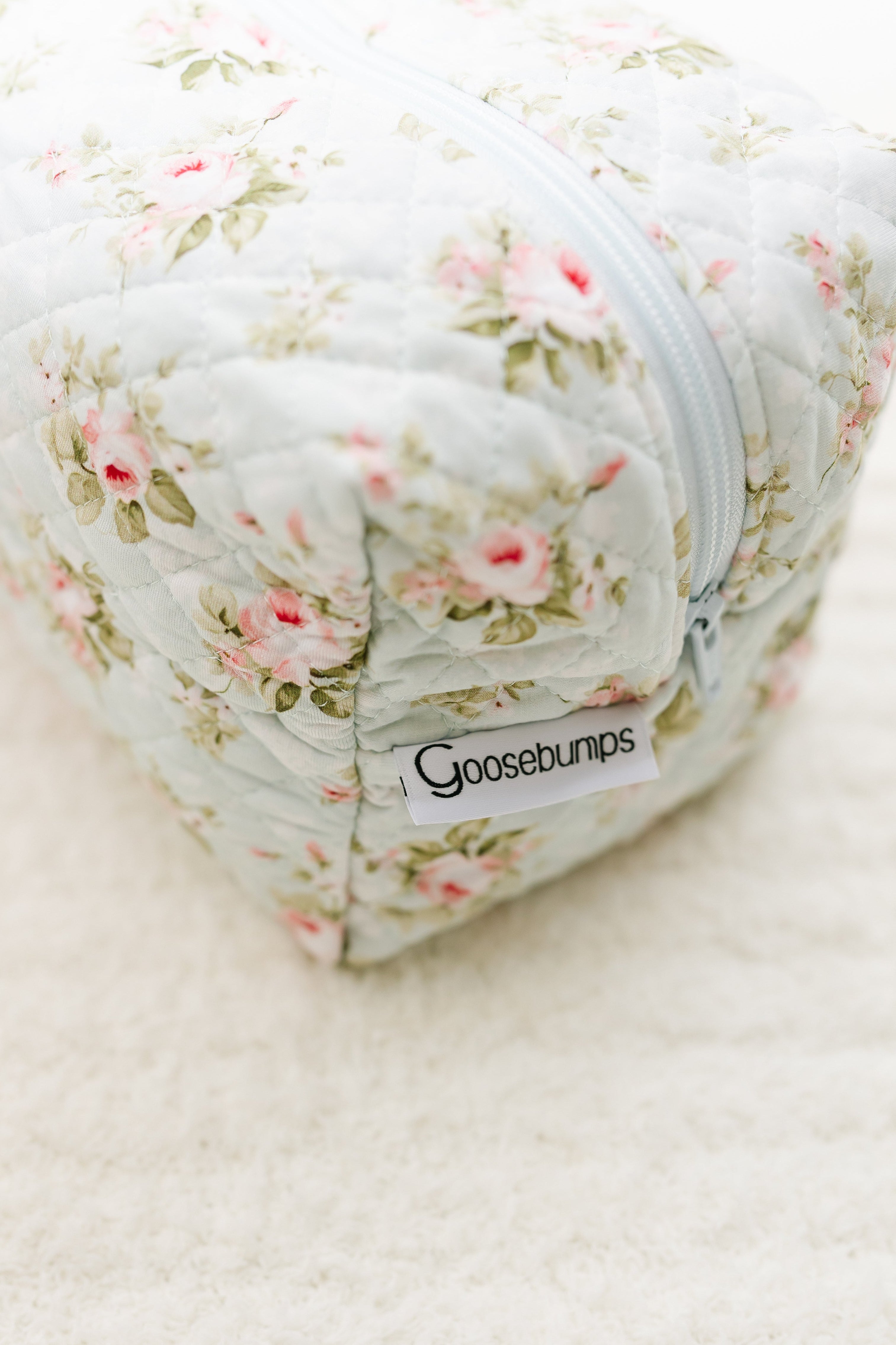A close-up of the "mint to be / travel bag," featuring quilted floral fabric with pink and green flowers, a white zipper, and a front label. The bag rests on a soft white surface.