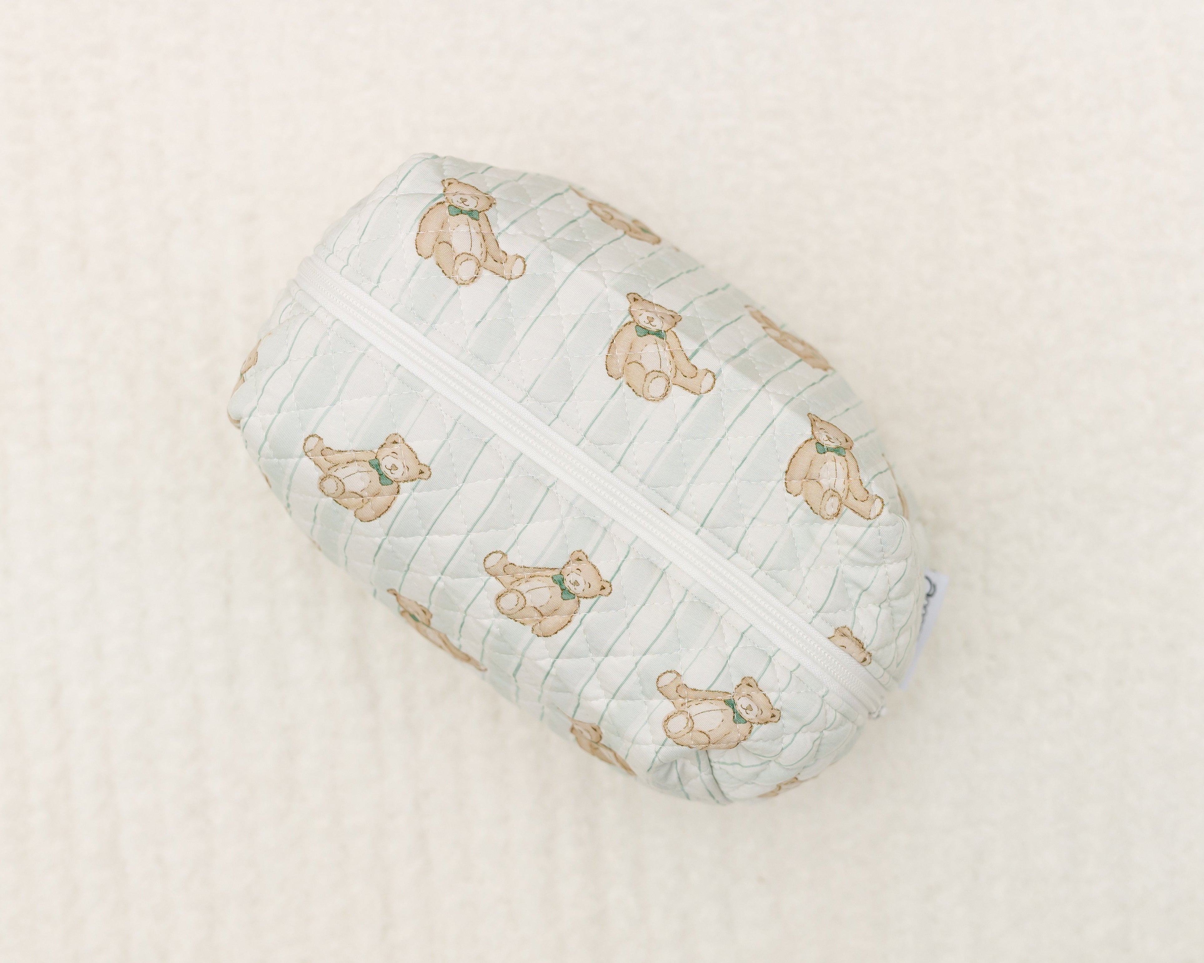 The teddy / travel bag is a small, quilted zipper pouch featuring light brown teddy bears with blue bows, ideal as a cute travel bag. It's shown on a soft, cream-colored textured surface.