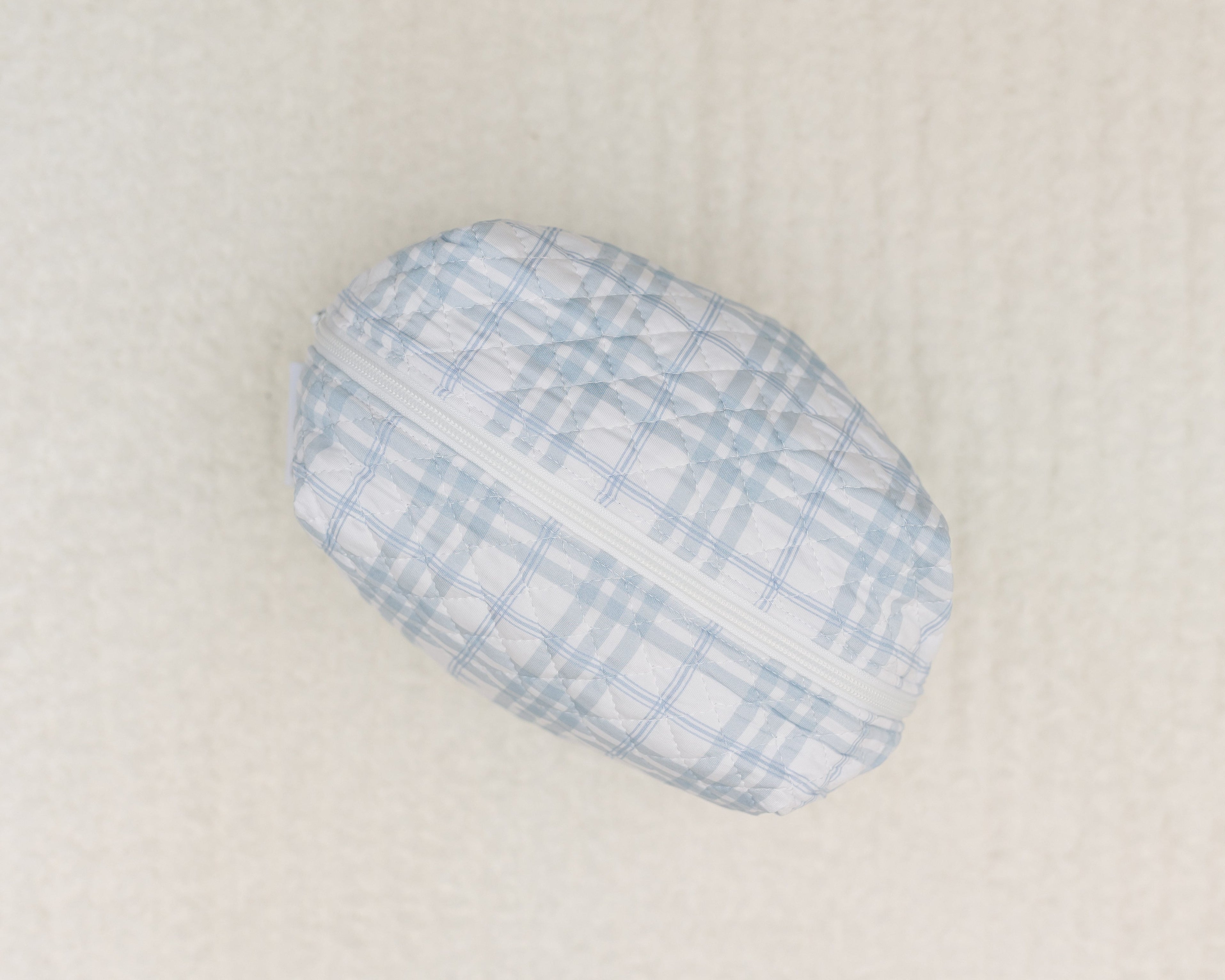 The luna travel bag is a small, oval-shaped bag featuring a light blue and white plaid print and a white zipper, shown resting on a soft cream-colored surface.