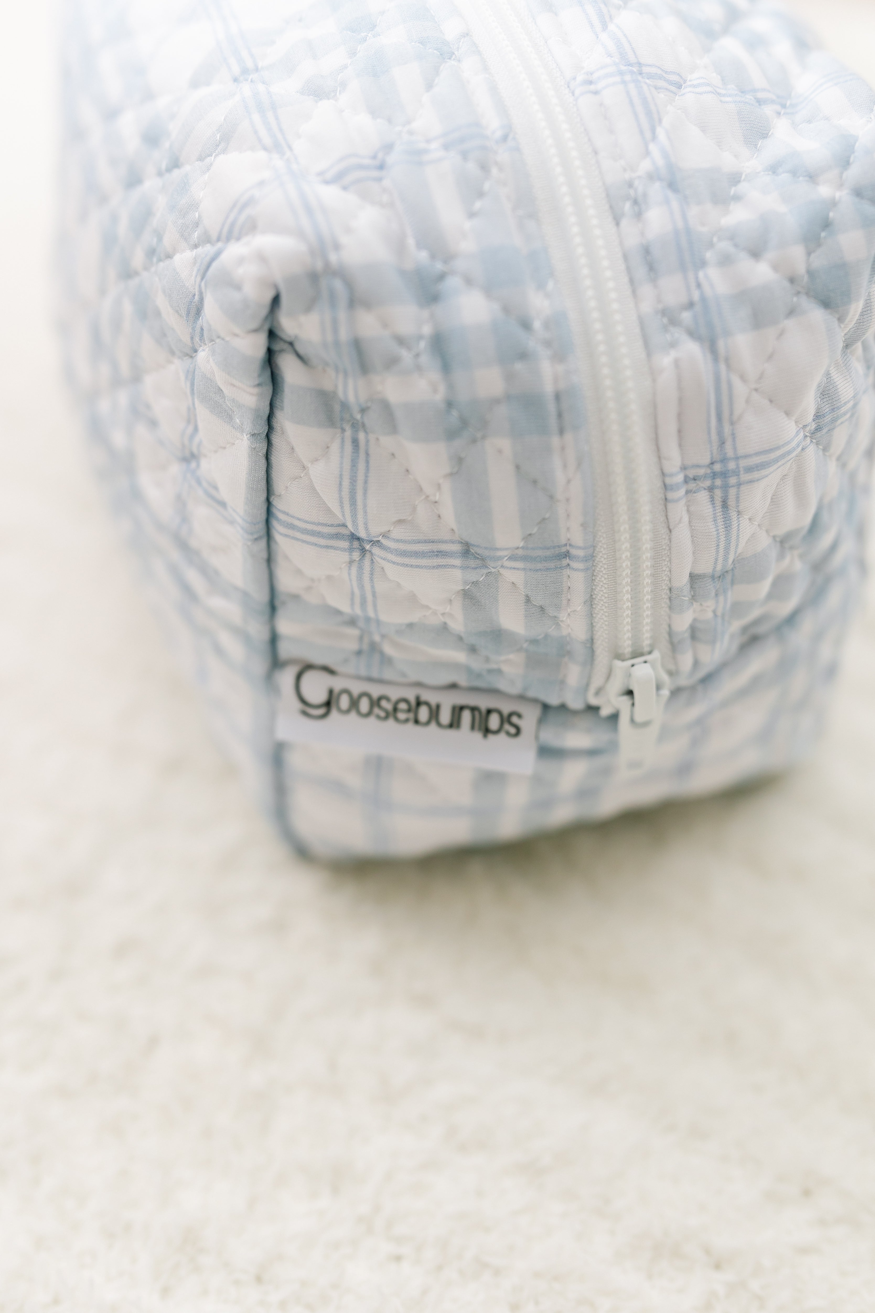 A close-up of the luna travel bag featuring a blue and white plaid print, quilted detailing, a white zipper, and a Goosebumps label, placed on a soft, light-colored surface.