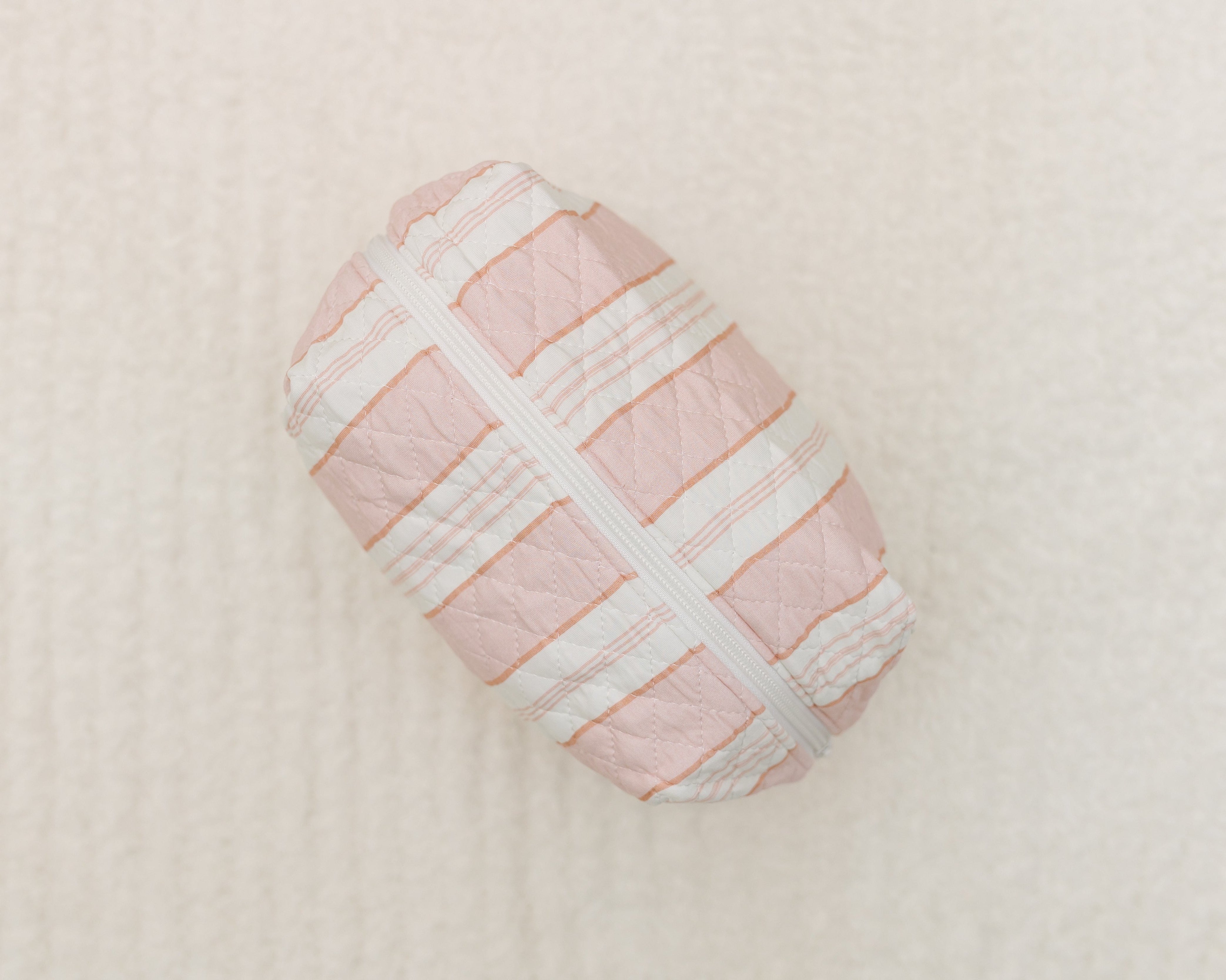 A small, quilted, rectangular pink pouch with white and peach stripes and a white zipper rests on a soft white surface—perfect as the lover girl / travel bag for carrying your essentials in style.