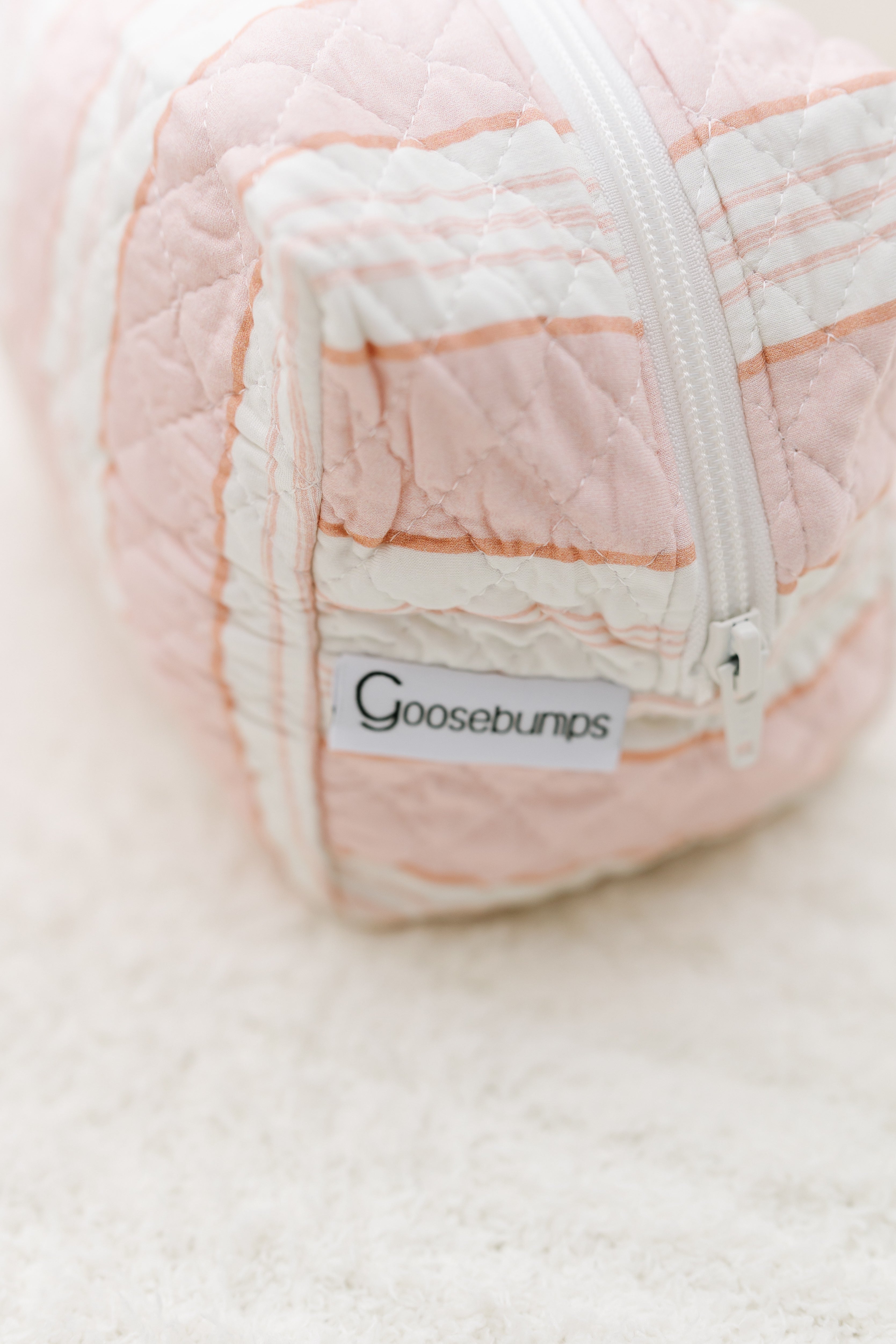 A close-up of the lover girl / travel bag, a pink and white quilted travel bag with a zippered top, set on a soft cream-colored textured surface.