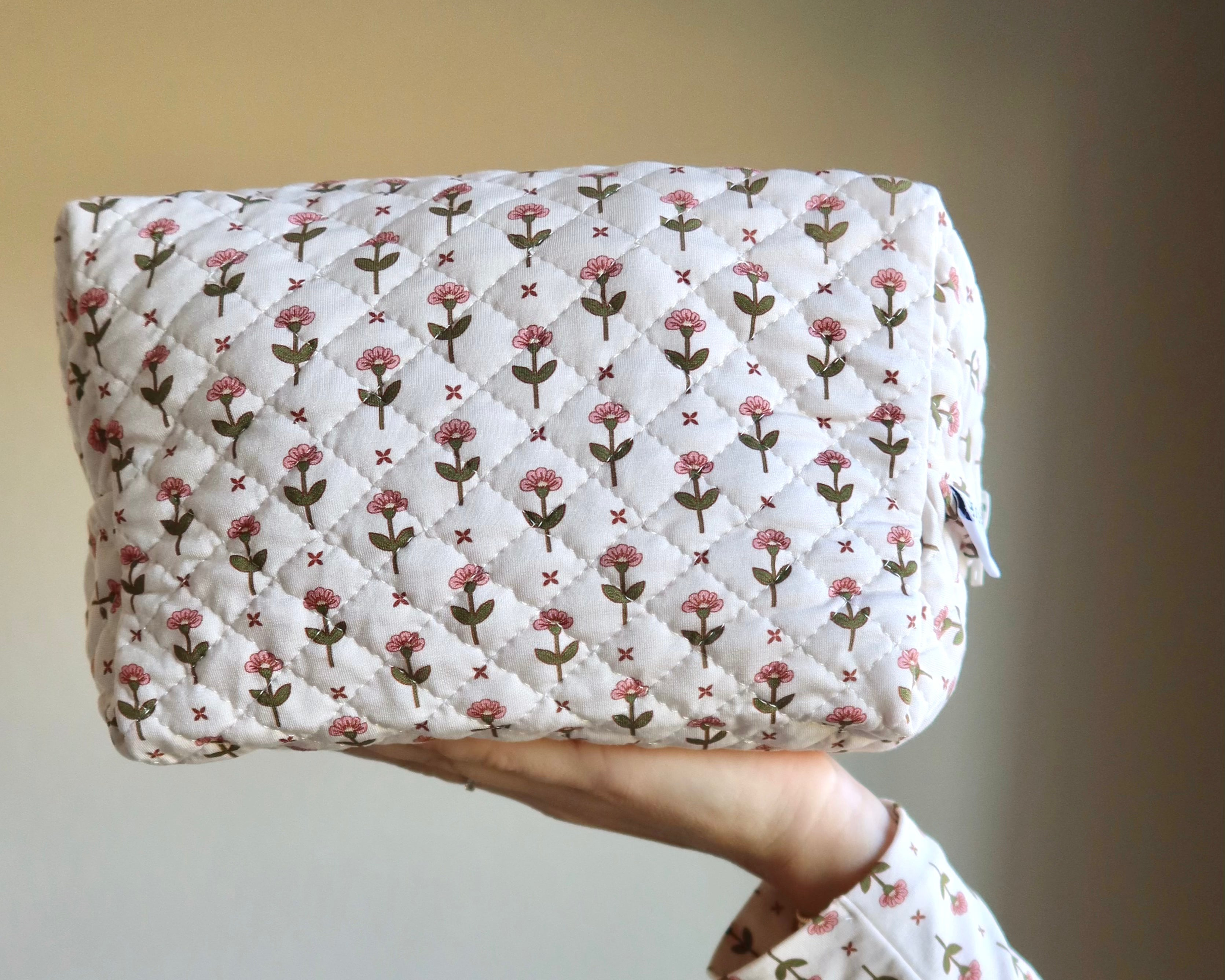 A hand holds the tulip / travel bag, featuring a white background with a repeating pattern of small pink flowers and green stems. The matching sleeve adds to the chic travel accessories look against the softly lit backdrop.