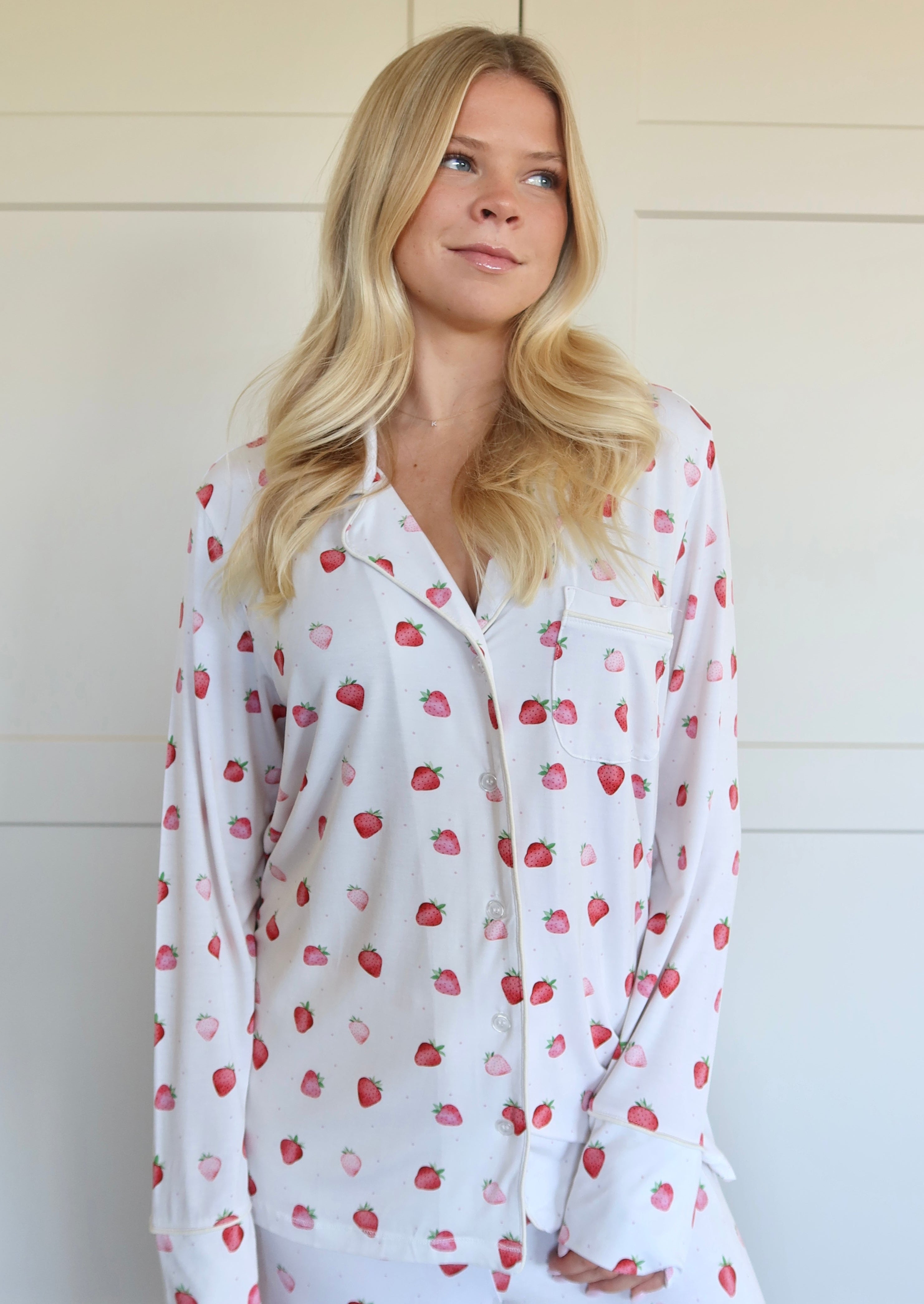 A woman with long blonde hair wears the strawberry shortcake long sleeve pajama set, featuring a relaxed fit and small red strawberry pattern, as she stands before a light-colored paneled wall.
