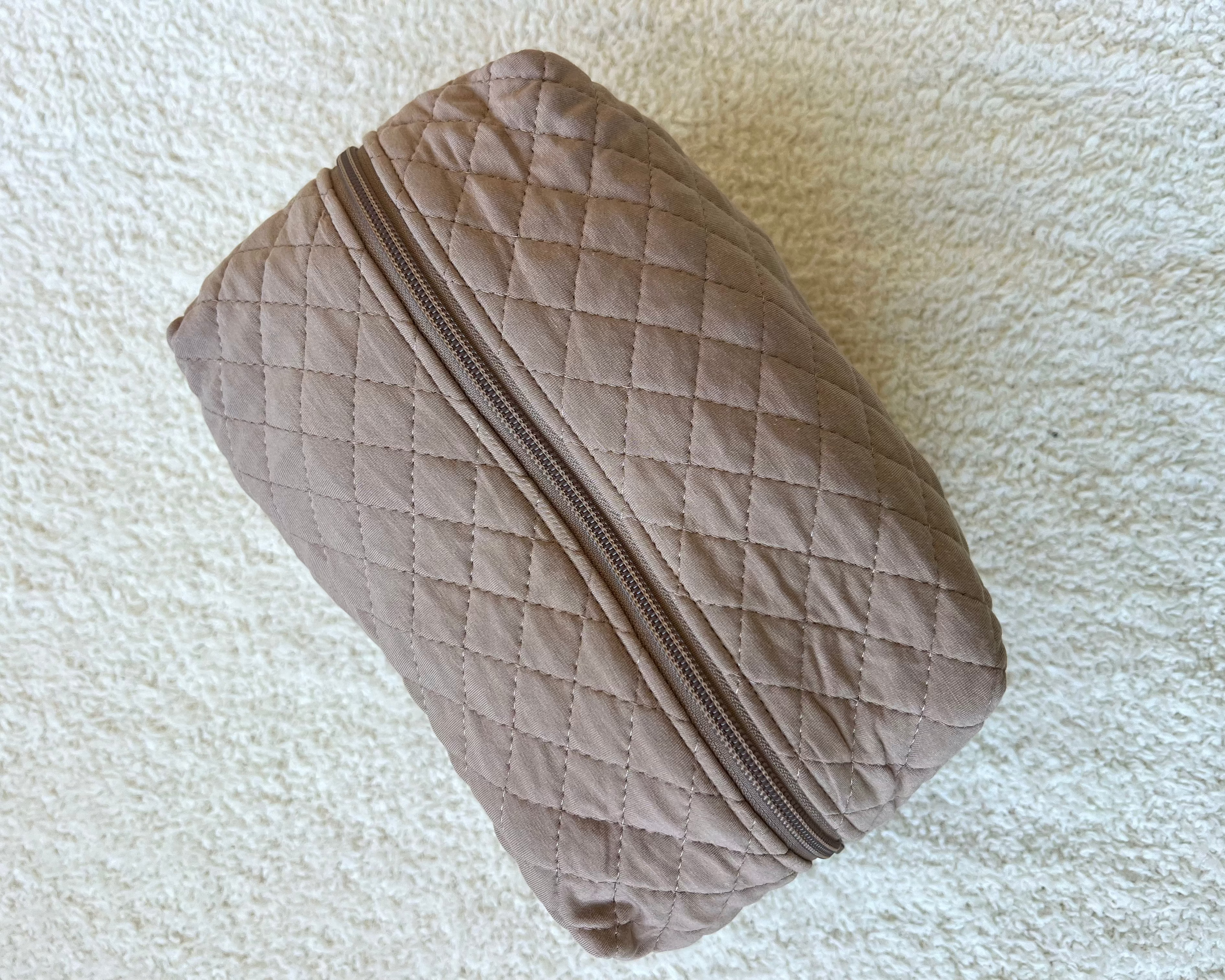 A gingerbread travel bag made of beige quilted fabric with a zipper rests on a cream-colored textured surface.