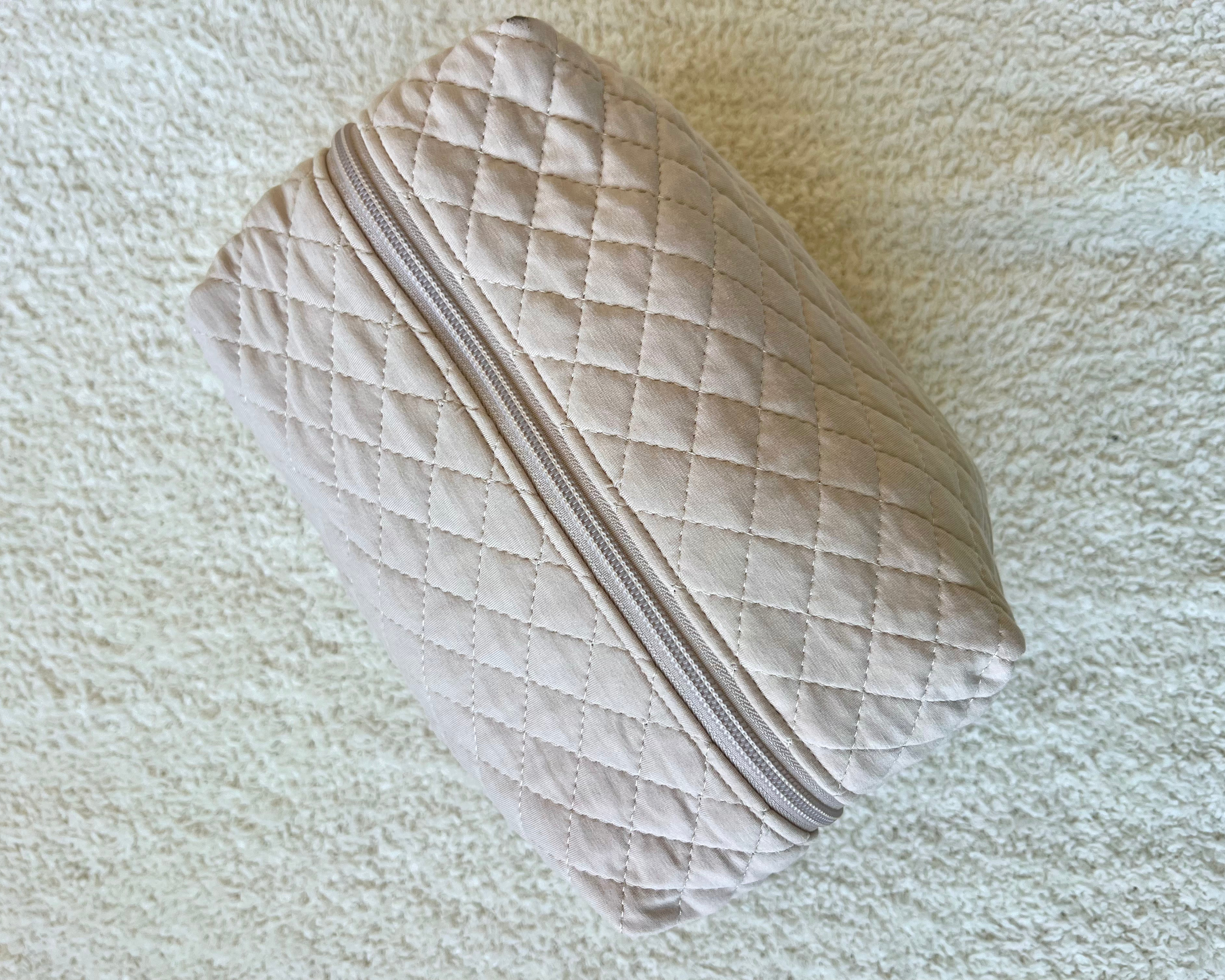 The vanilla cream travel bag, crafted from beige quilted bamboo viscose with a zipper, rests on a light textured surface—an elegant and eco-friendly addition to your travel accessories.