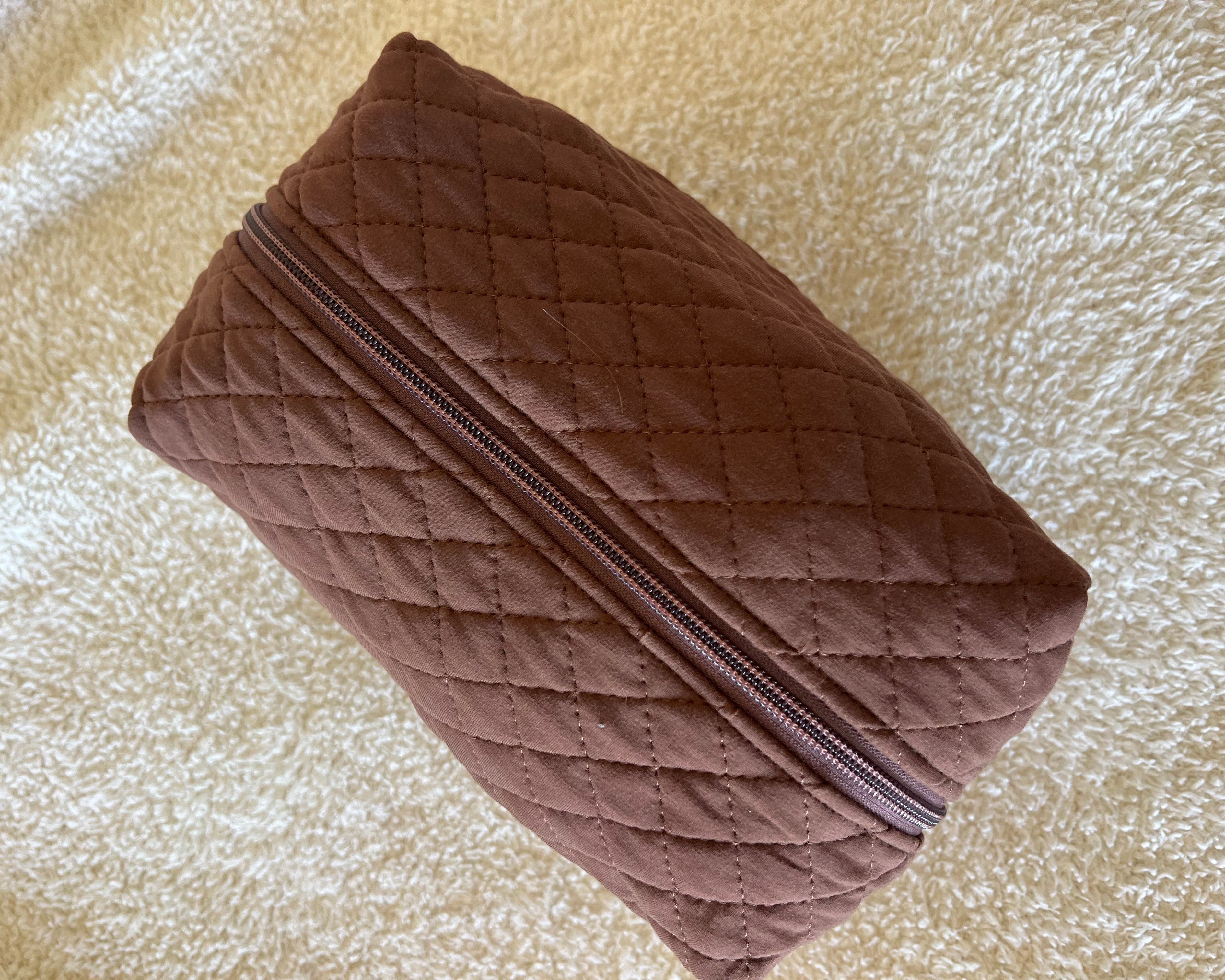 A chocolate-colored quilted travel bag with a zipper rests on a textured cream surface.