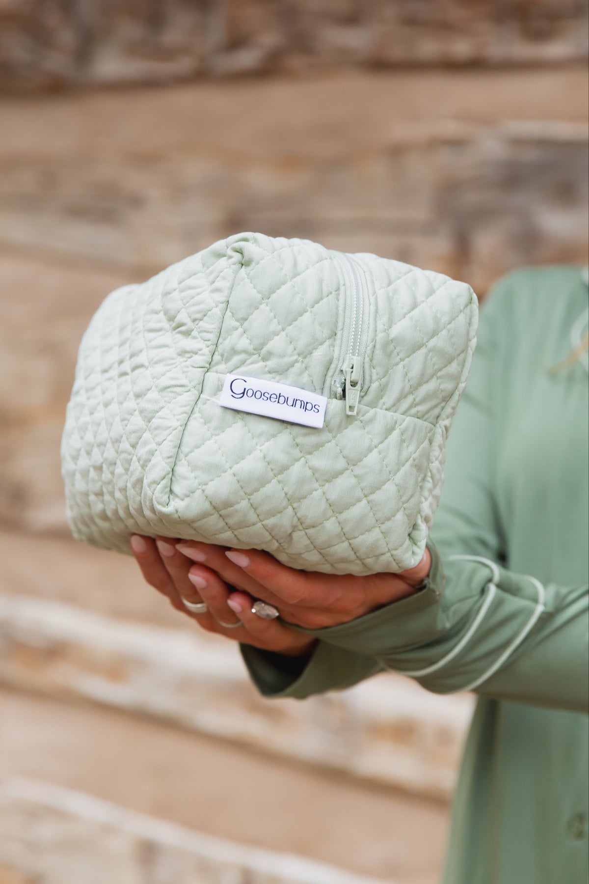 A person holds a solid sage green, quilted frosted fir travel bag with a zippered top and a Goosebumps brand tag.