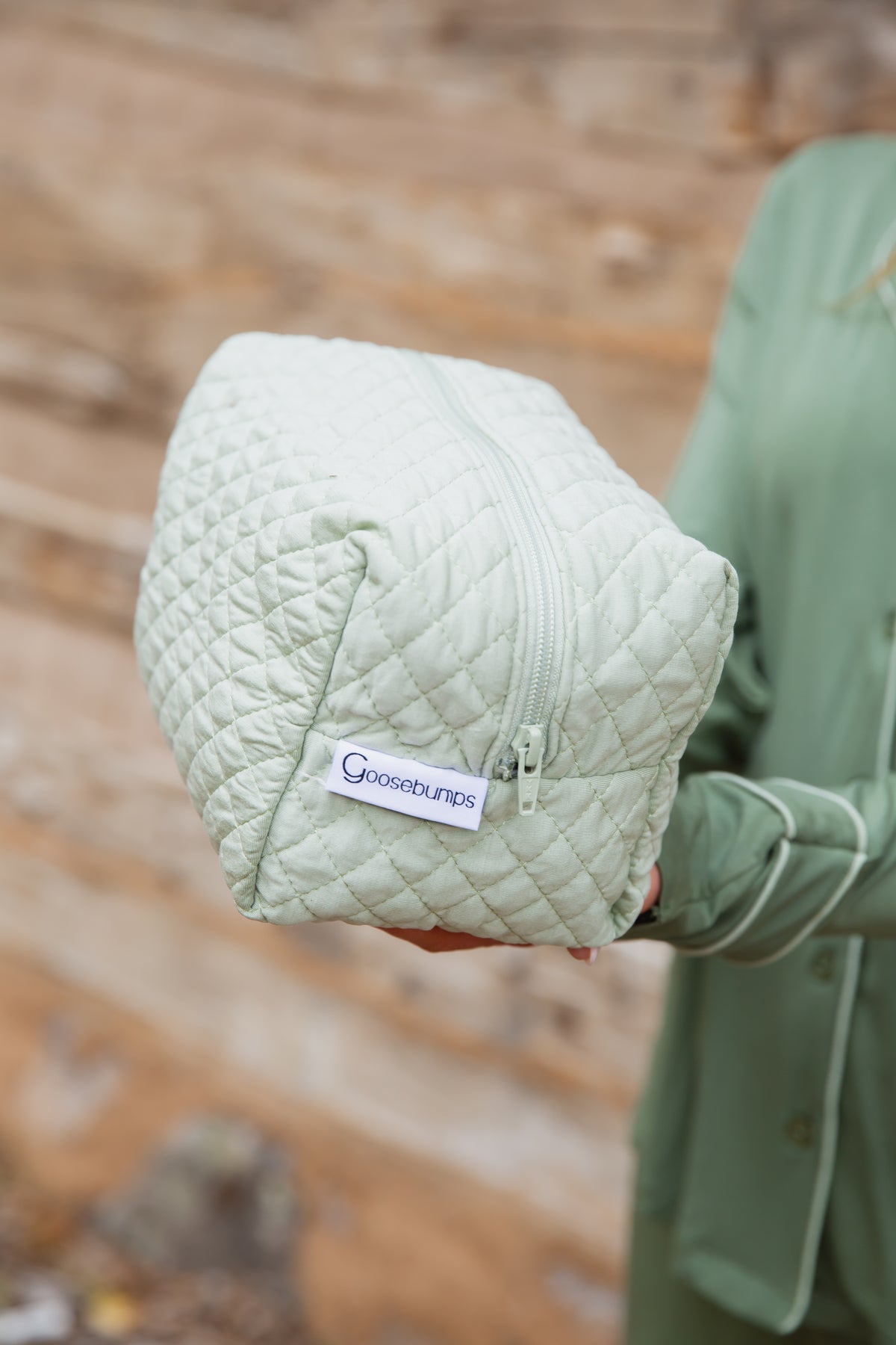 A person holds an 8″x6″x6″ frosted fir quilted travel bag with a zipper closure and a branded tab.
