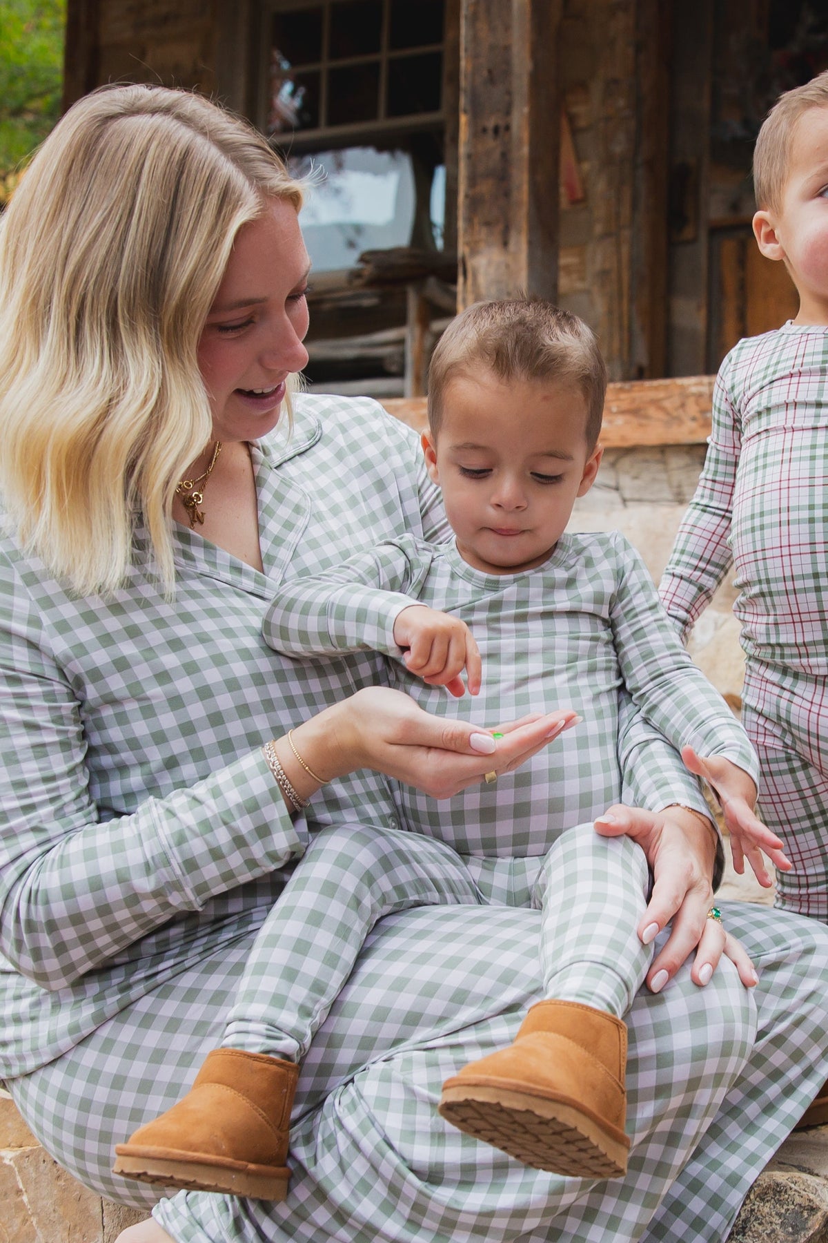 A woman and two young boys wear matching green and ivory gingham evergreen lane / two piece skinny bamboo pajamas, with the woman holding a small item for one of the boys.