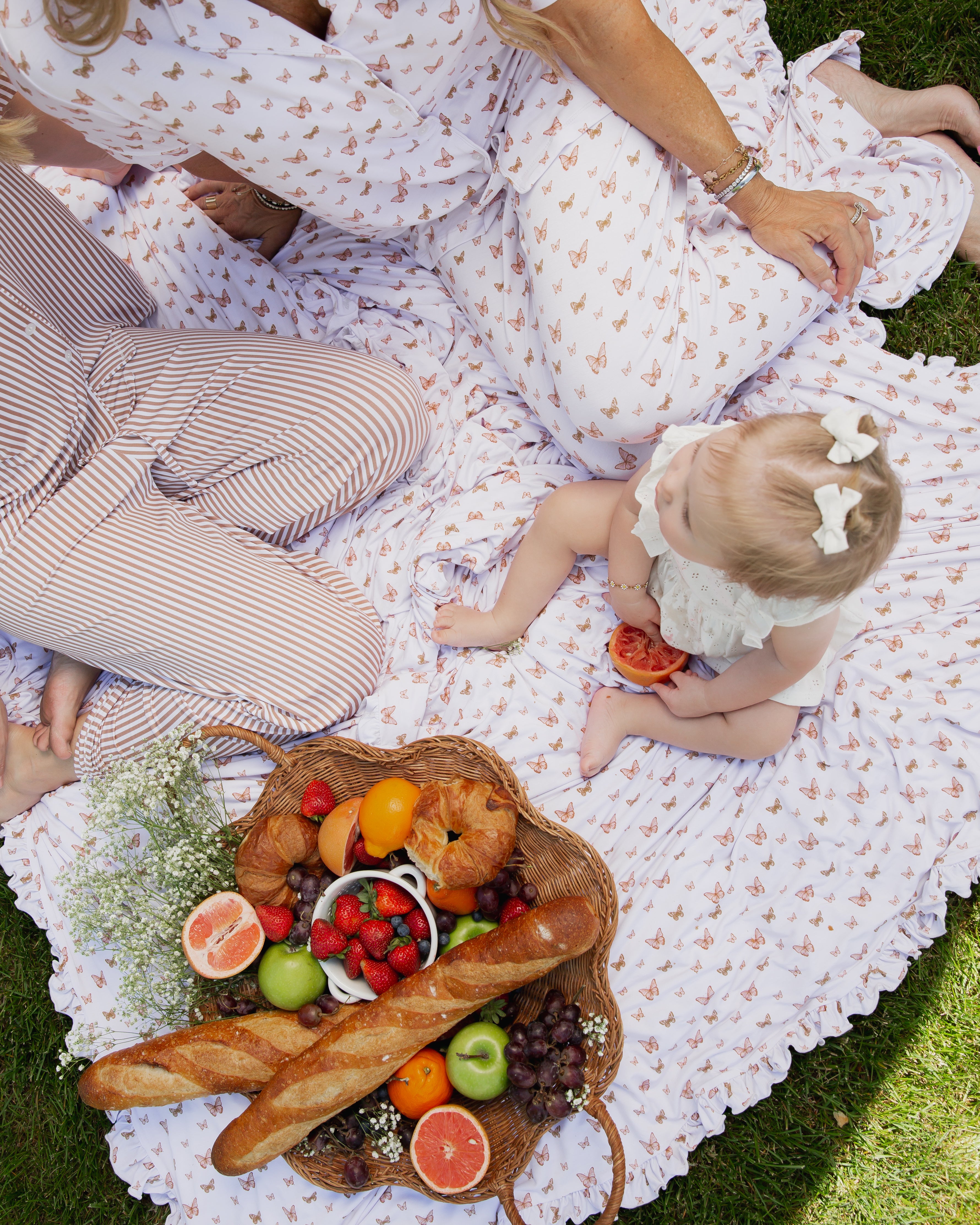 A baby with a white bow sits outdoors on a patterned blanket with two adults, wrapped in the soft "flutter / ruffle" bamboo swaddle, surrounded by a picnic of fruit, croissants, grapes, strawberries, and baguettes in a basket.