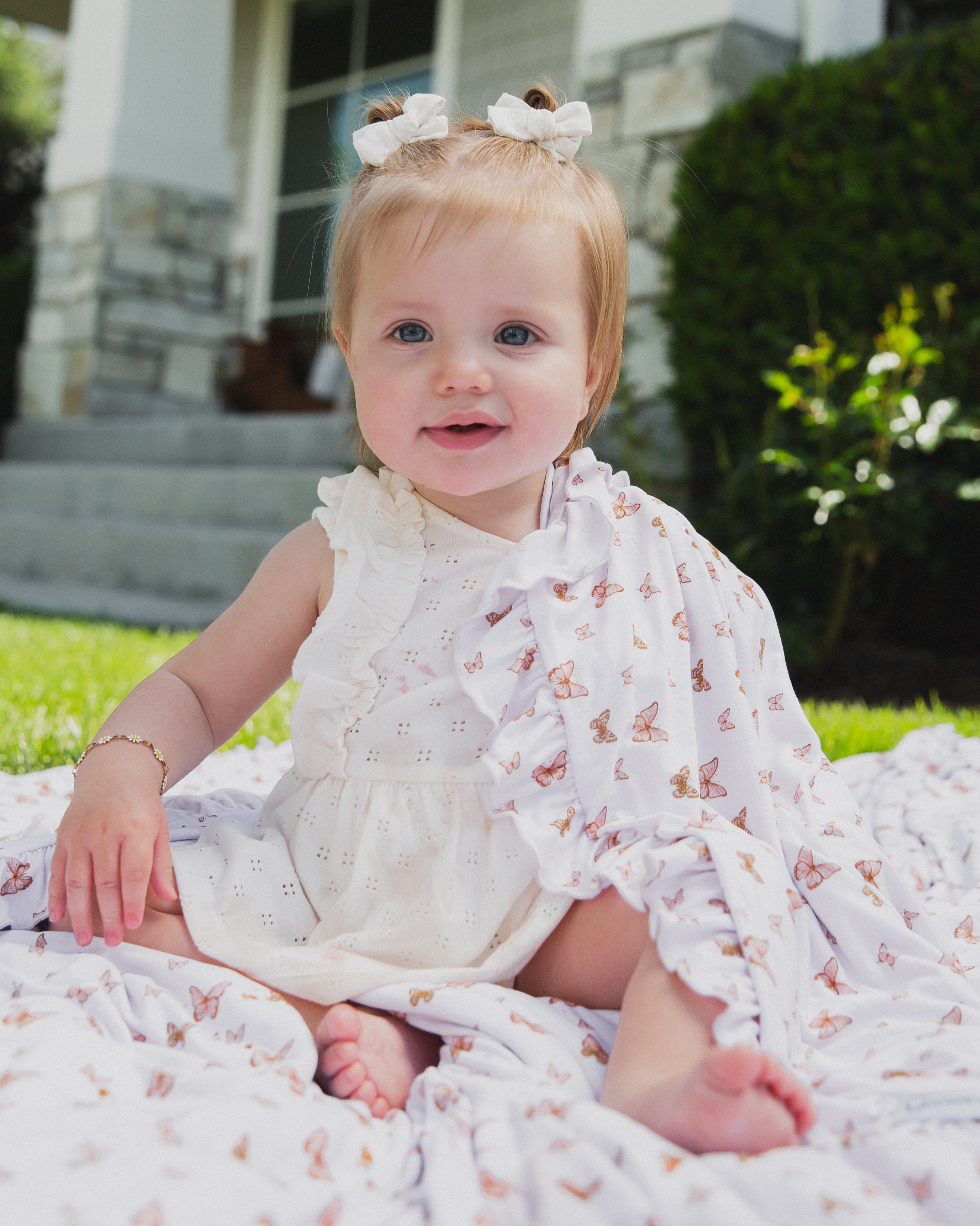 A smiling baby with light hair sits on a patterned blanket outdoors, wearing the flutter / ruffle white dress. Hair bows, green grass, and a house in the background highlight the comfort of this cozy bamboo viscose style.