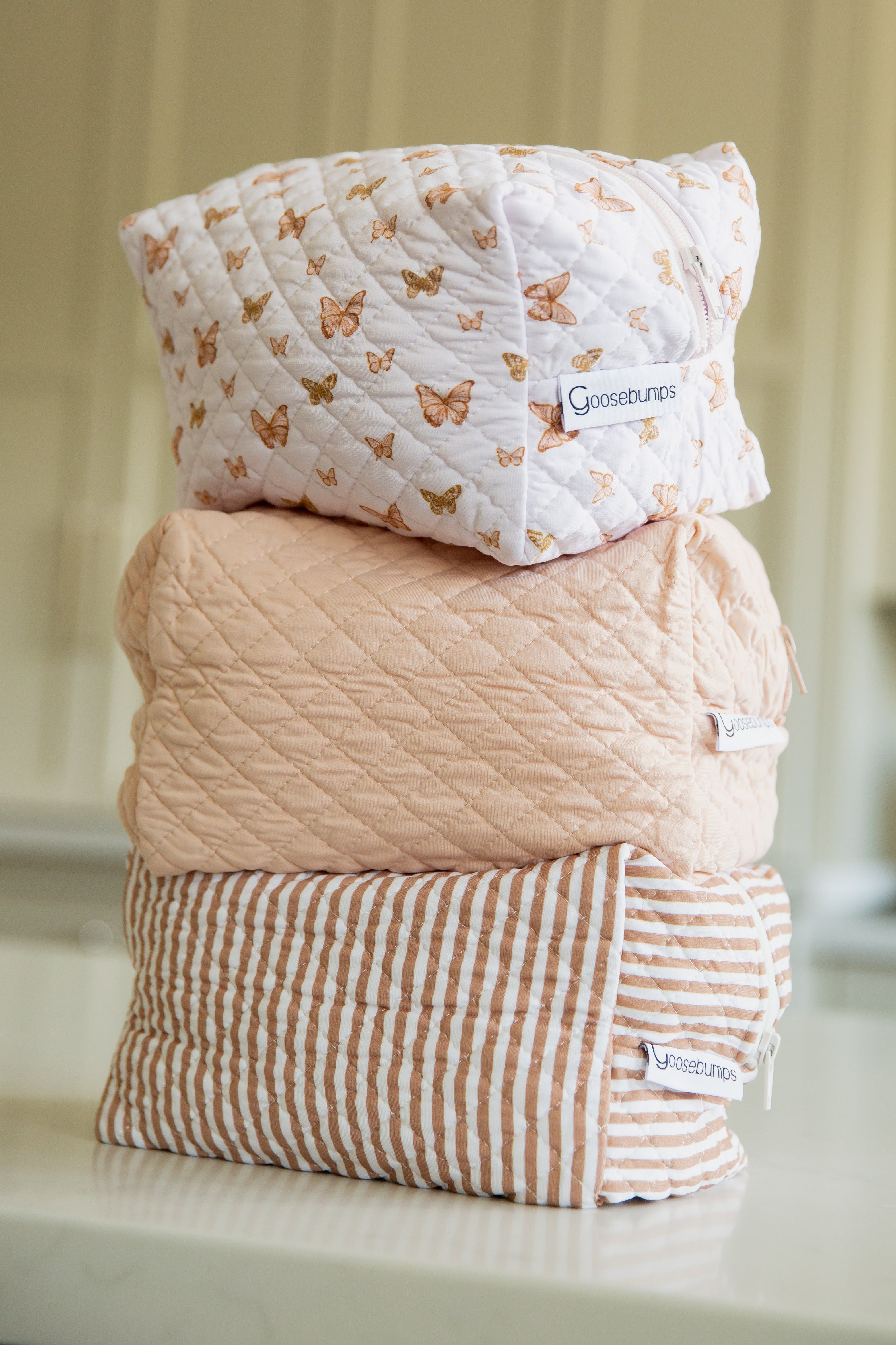 Three whisp/travel bags from the Lisa Wilson Goosebumps Collection are stacked: the top is white with a butterfly print, the middle is solid peach, and the bottom is peach and white striped, each featuring Goosebumps labels.