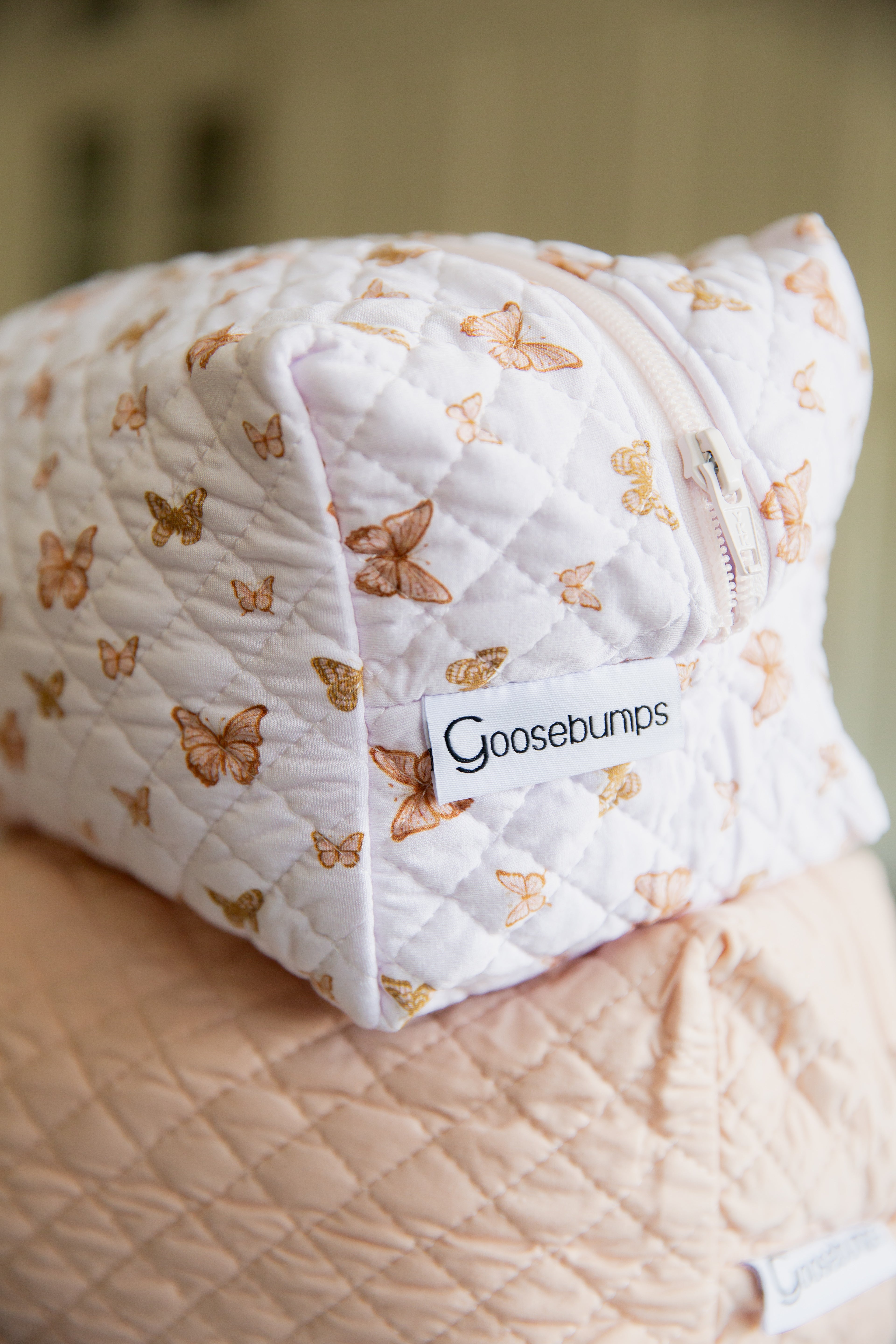 The flutter / travel bag from the Lisa Wilson Goosebumps Collection features a quilted white butterfly print, visible zipper, and signature label. Perfect for travel, it’s shown on a light peach quilted surface.