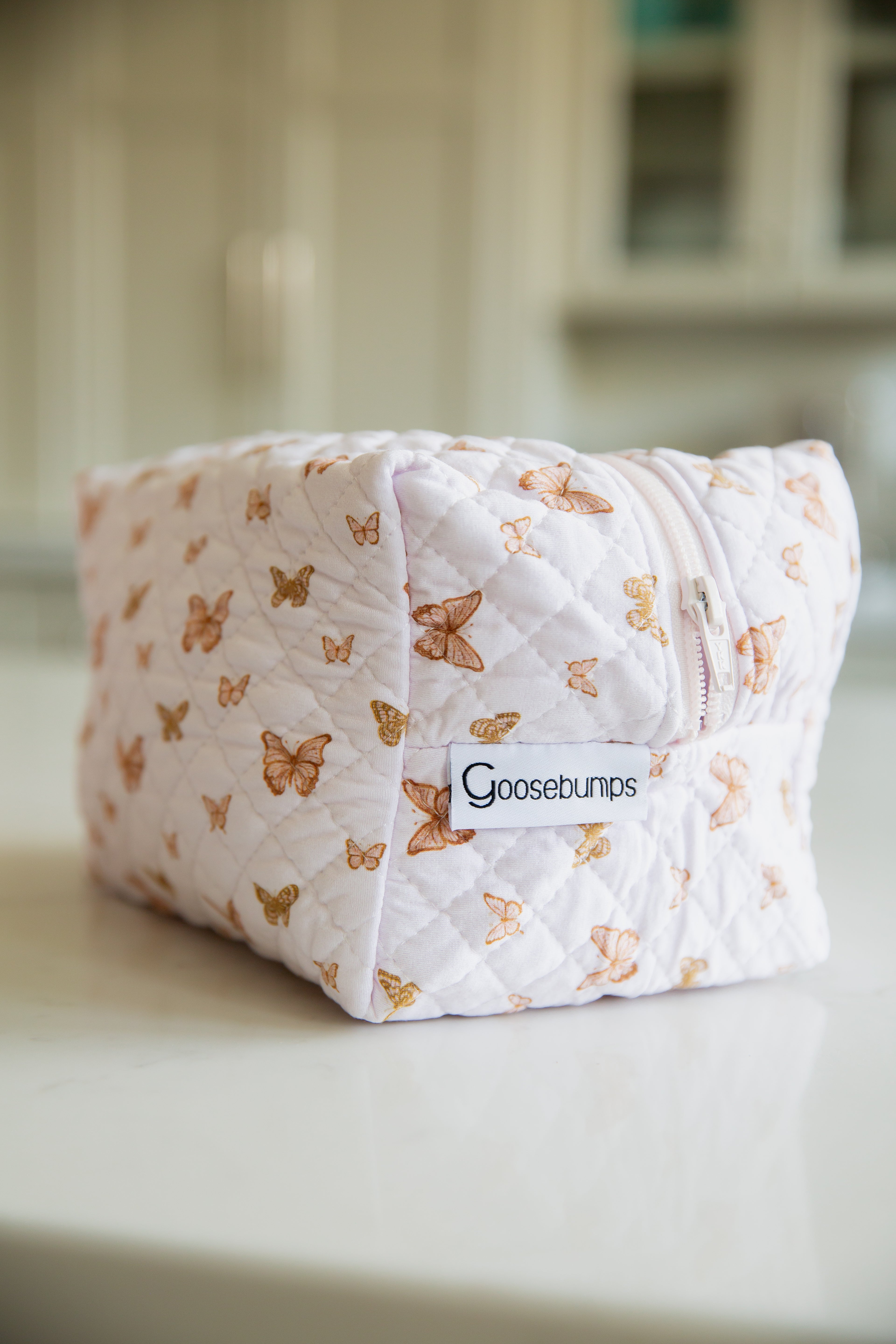 A quilted, rectangular flutter / travel bag with a pink and orange butterfly pattern and a zipper sits on a white surface. A label reads Goosebumps from the Lisa Wilson Goosebumps Collection; the background is softly blurred.