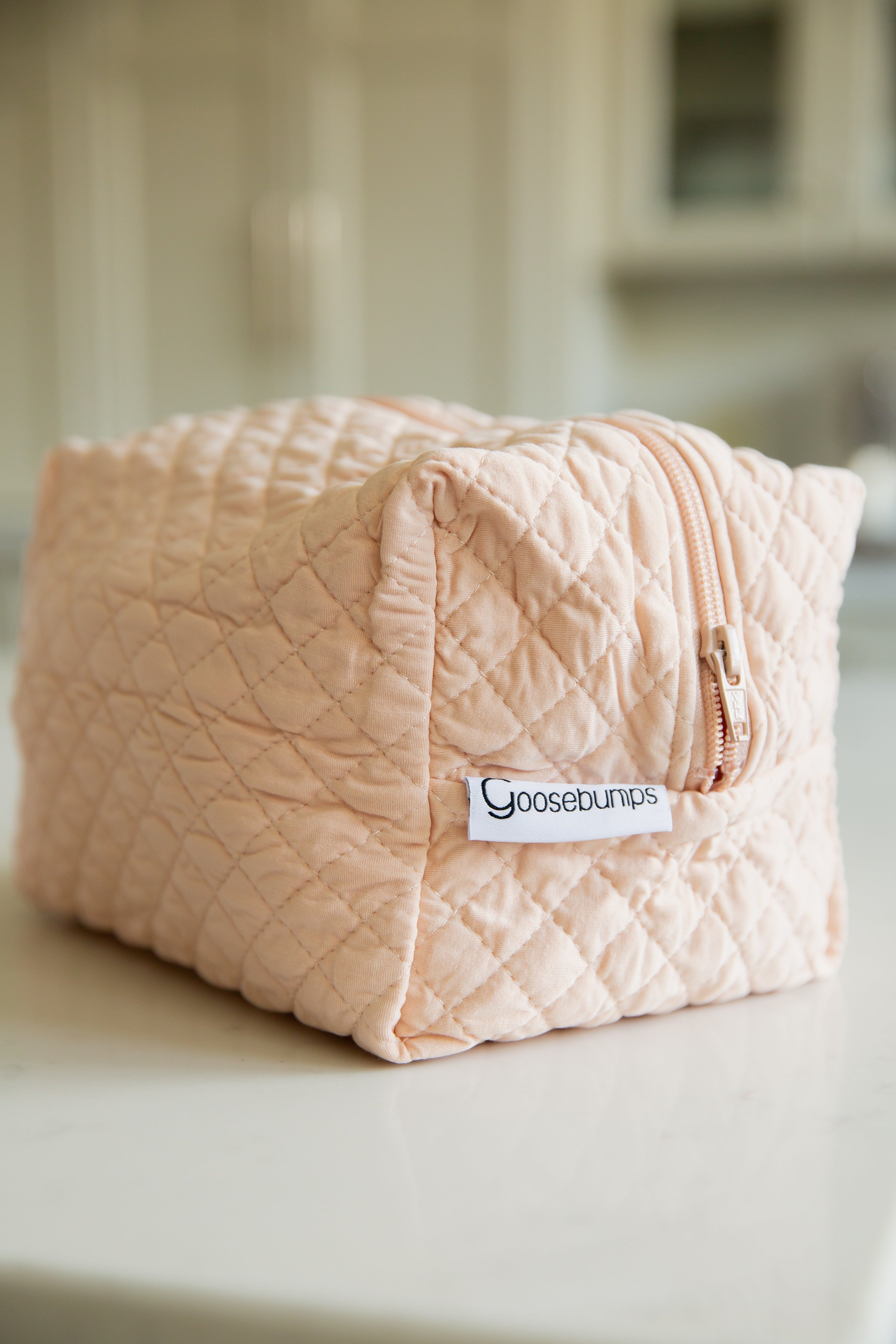 The whisp / travel bag from the Lisa Wilson Goosebumps Collection, a light pink quilted zipper pouch, rests on a white countertop—ideal for travel—with a blurred kitchen background.