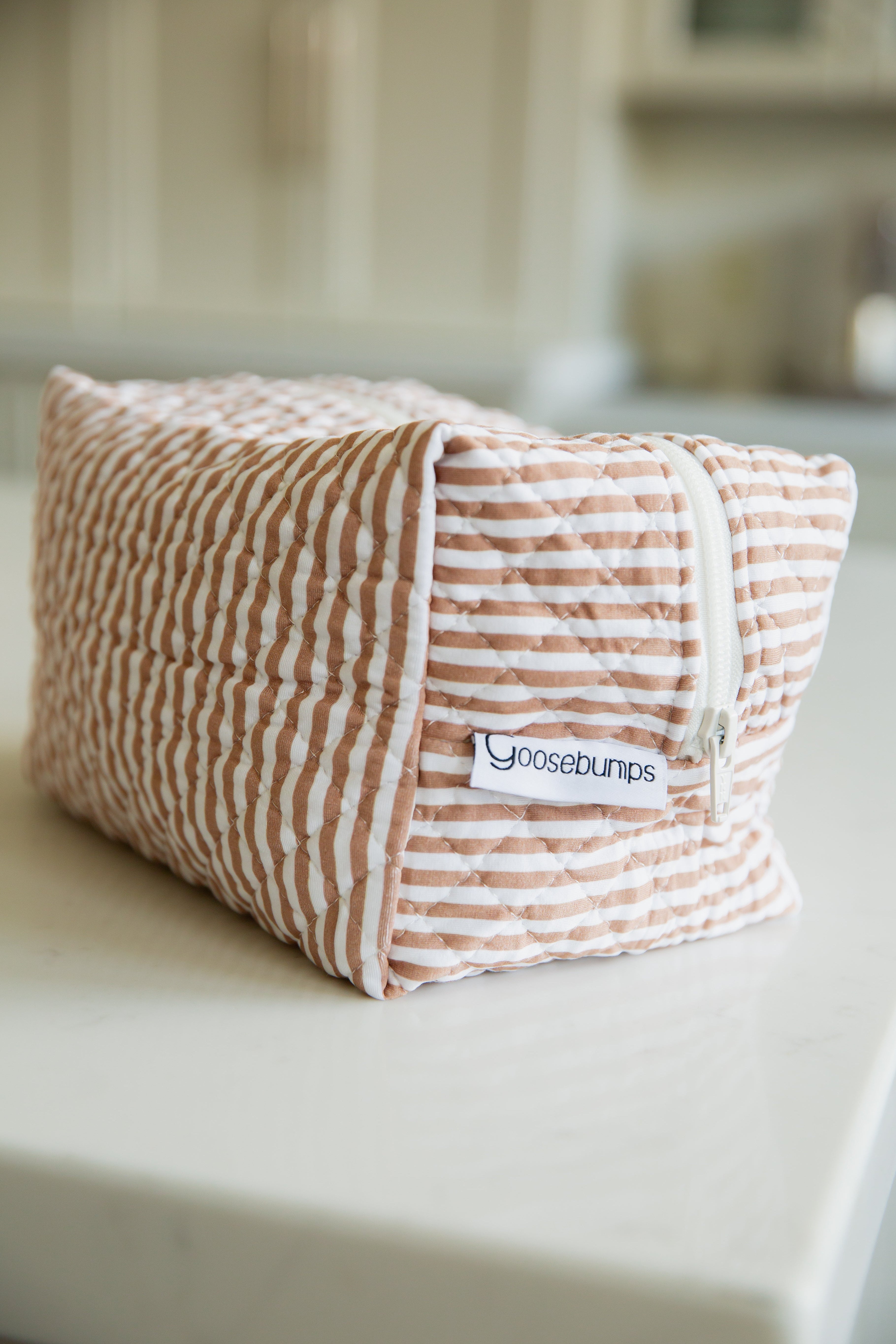 A rectangular, quilted drift / travel bag with a white zipper and peach and white stripes sits on a white countertop, featuring a Goosebumps label from the Lisa Wilson Goosebumps Collection.