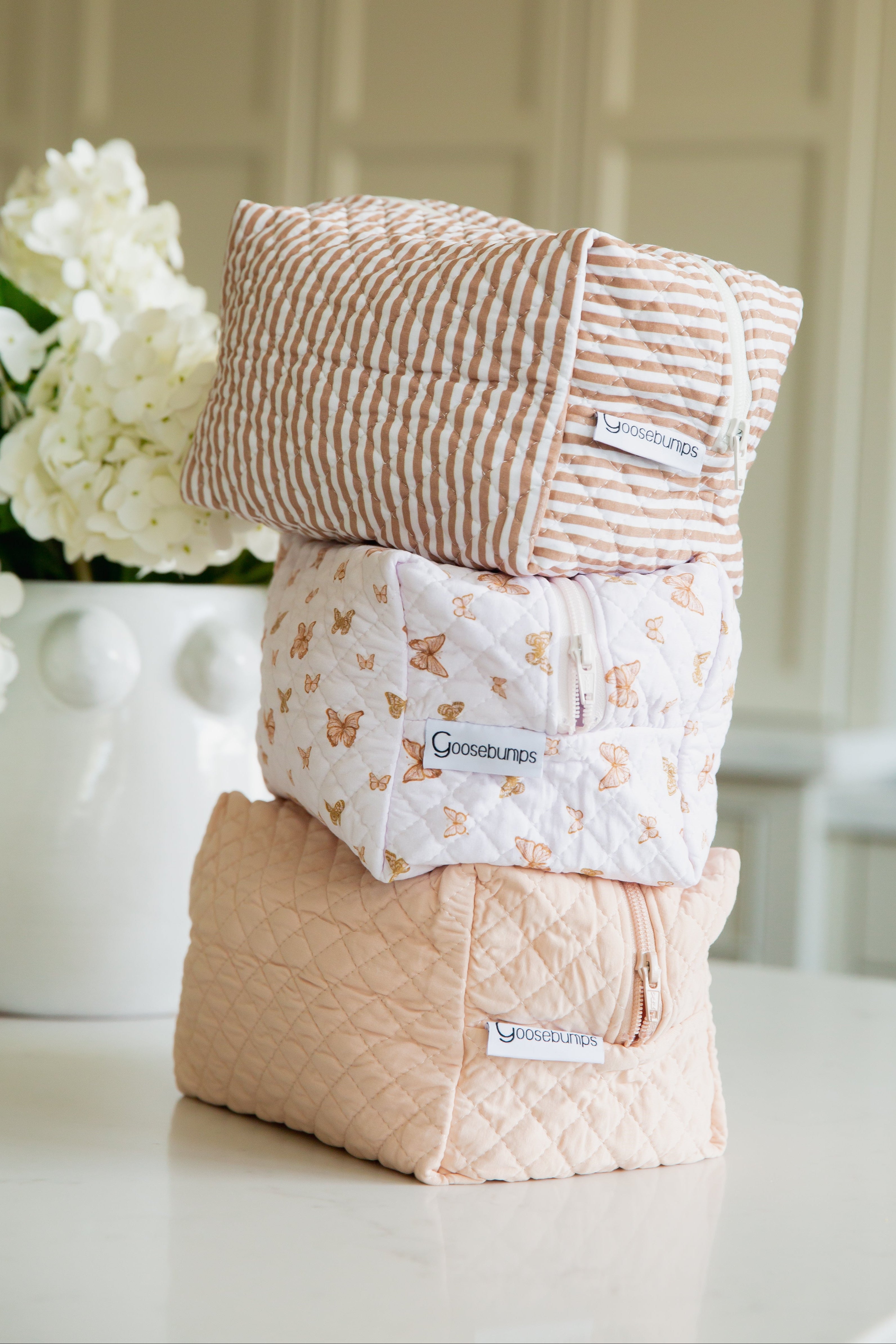 Three pastel quilted "drift / travel bag" toiletry bags are stacked on a white countertop. Each features unique striped, butterfly, or plain prints from the Lisa Wilson Goosebumps Collection, with white flowers in the background.