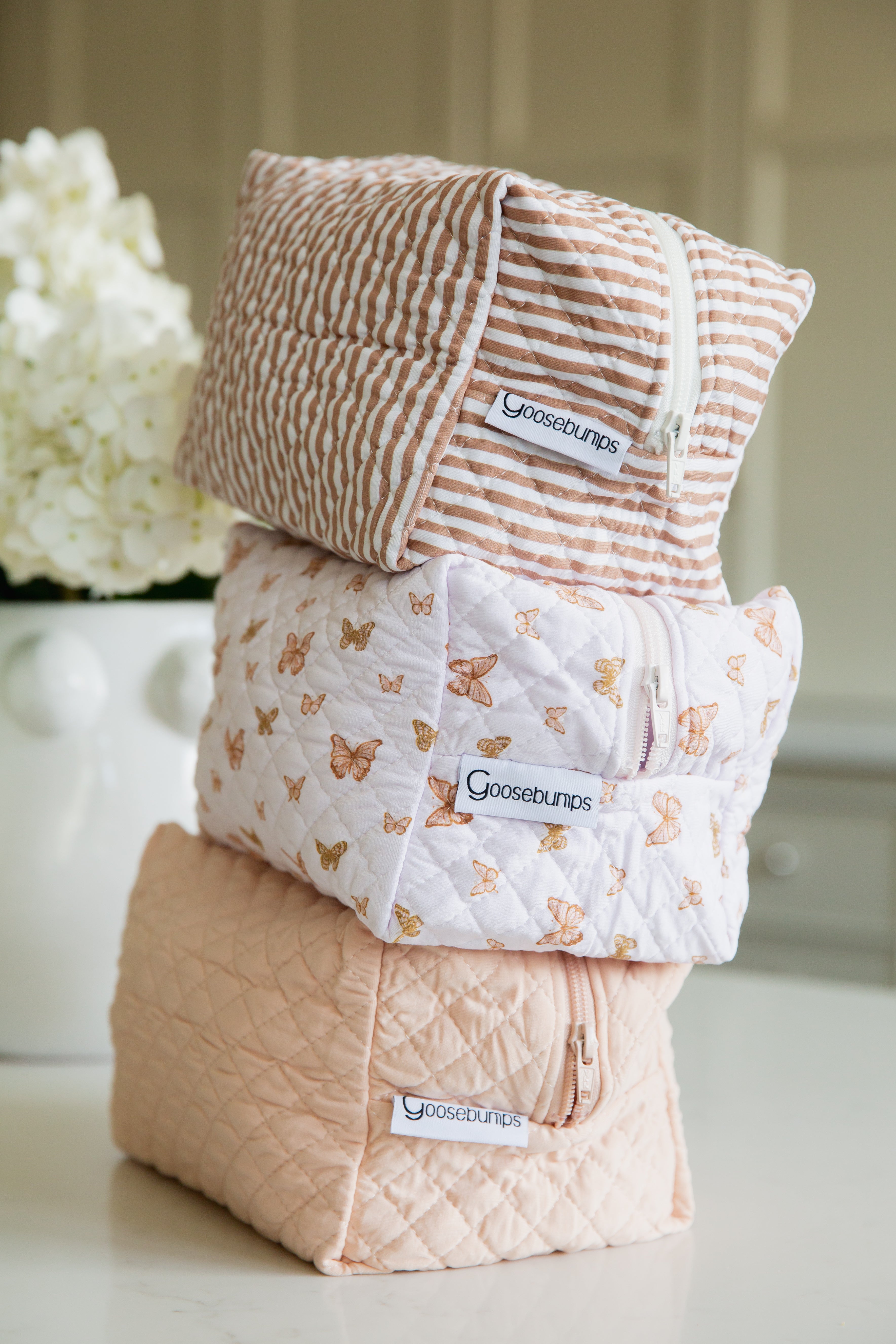 Three soft zippered pouches from the flutter / travel bag line are stacked: brown and white stripes on top, butterfly print in the middle, quilted pink on the bottom. A white vase with flowers is in the background.