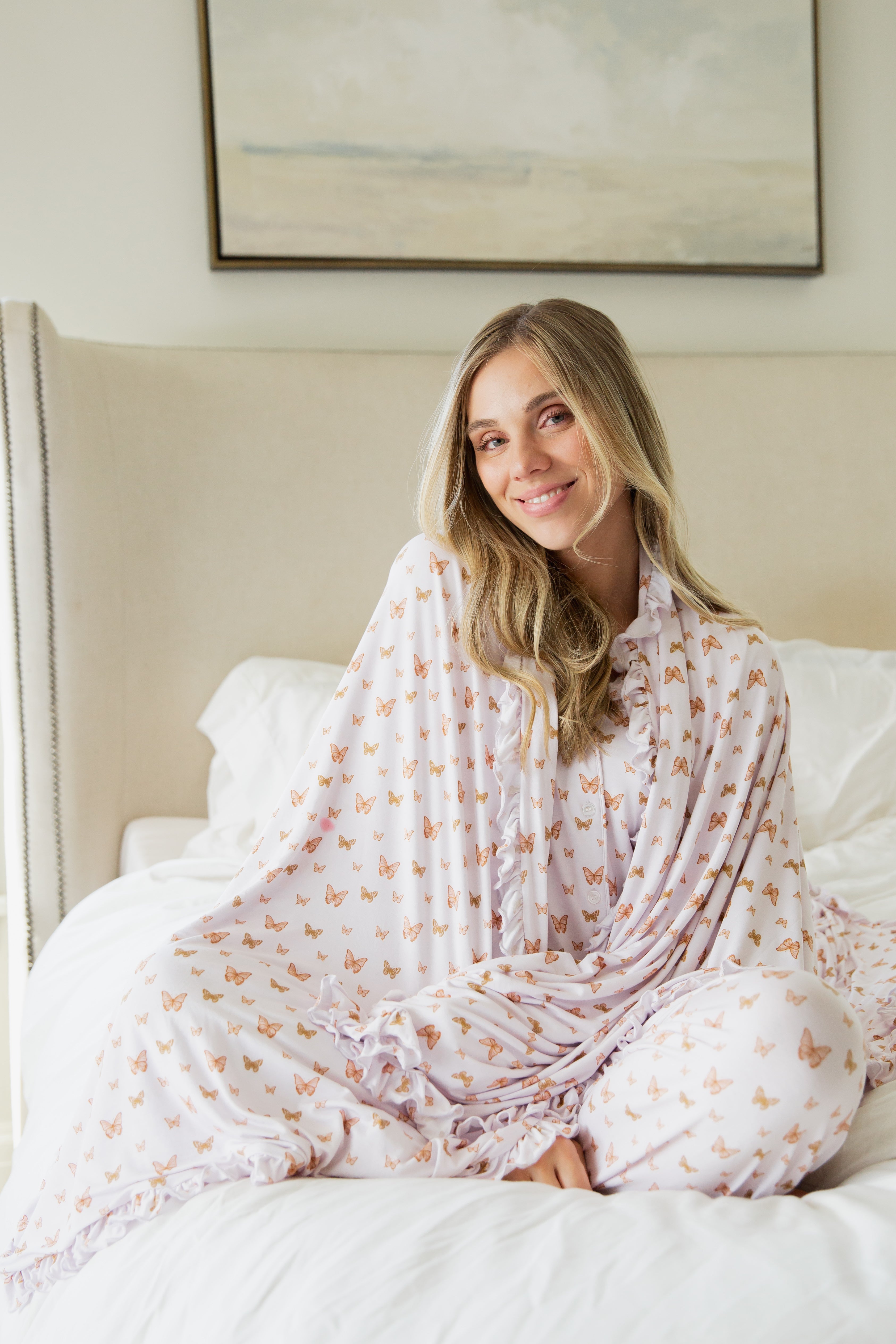 A woman with long blonde hair sits on a bed, smiling, wrapped in the flutter / ruffle adult swaddle featuring a butterfly pattern and delicate ruffle trim. The softly lit room has a beige headboard and a framed painting on the wall behind her.