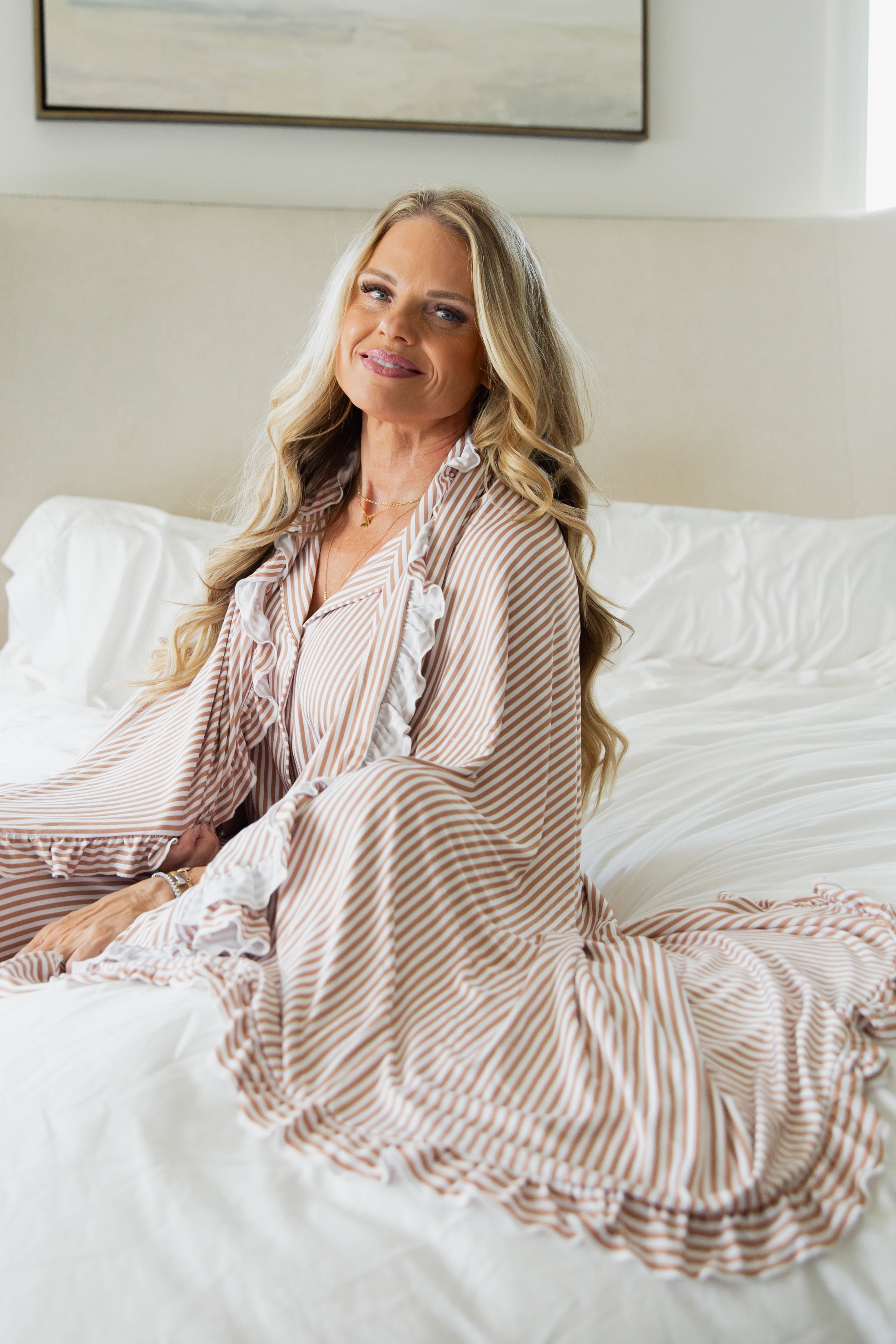 A woman with long blonde hair sits on a bed, smiling at the camera. She is wearing the drift / ruffle striped robe from the Goosebumps Collection, surrounded by white bedding in a softly lit bedroom.