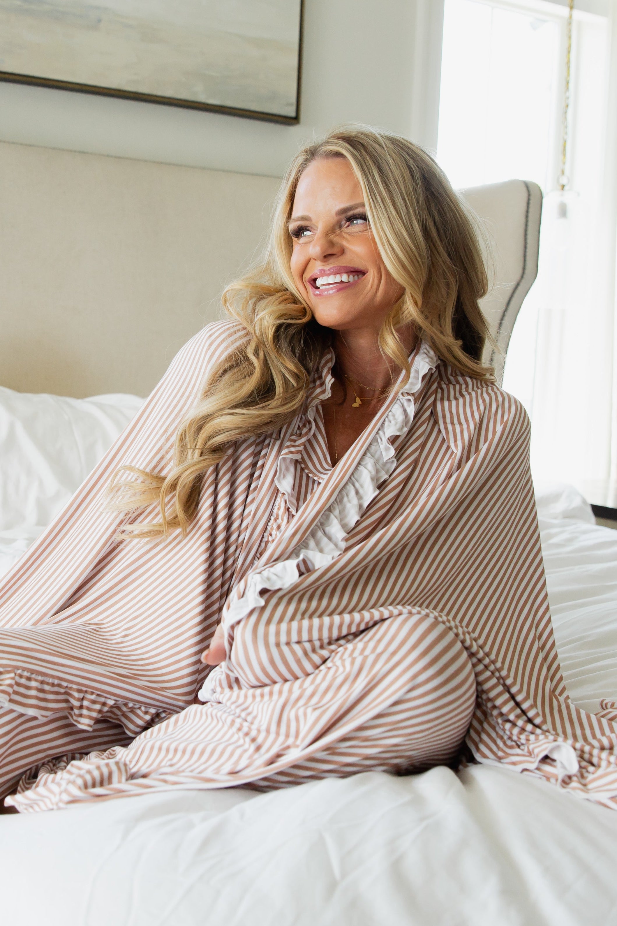 A smiling woman with long blonde hair sits on a bed, wearing the drift / ruffle loungewear set. She looks to the side in a cozy, sunlit bedroom filled with soft, natural light.