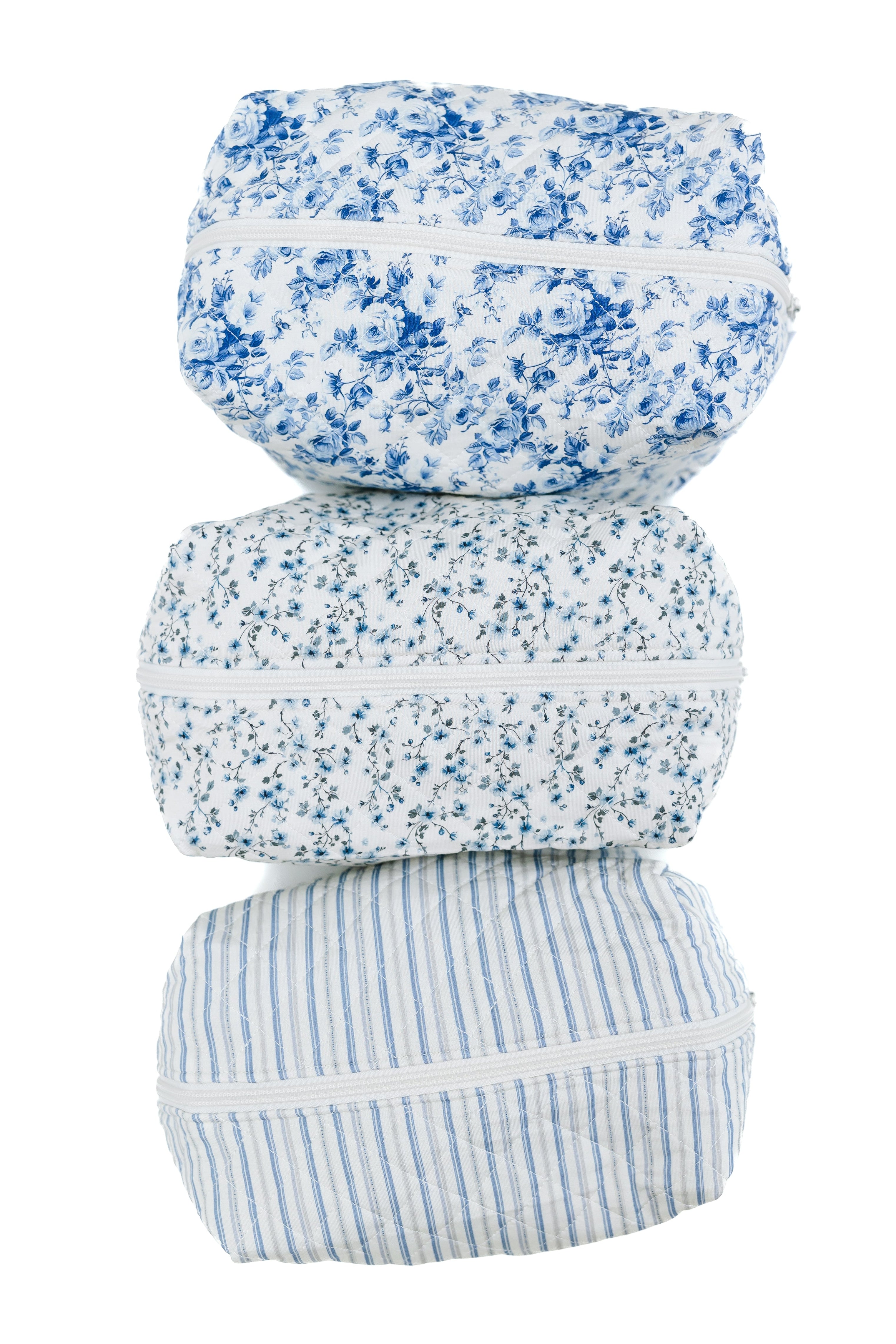 Three fabric pouches stacked vertically as the beach house / travel bag: top with blue floral, middle with small blue floral, bottom with blue stripes; all feature white zippers and backgrounds—ideal for travel accessories.