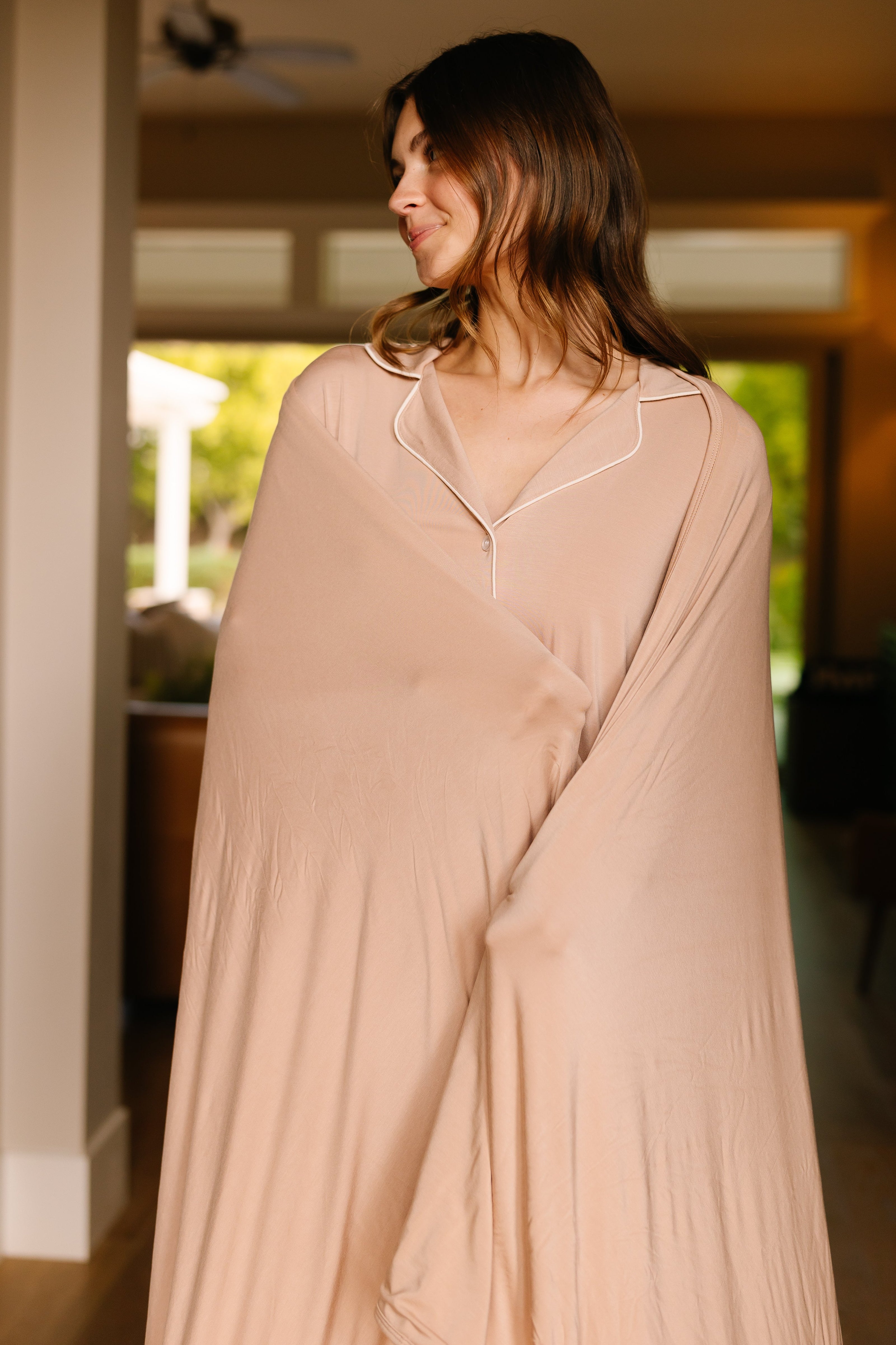 A woman with long brown hair stands indoors, wrapped in the luxurious almond blanket made from viscose from bamboo. She smiles softly as natural light streams through the windows, creating a warm and cozy atmosphere.