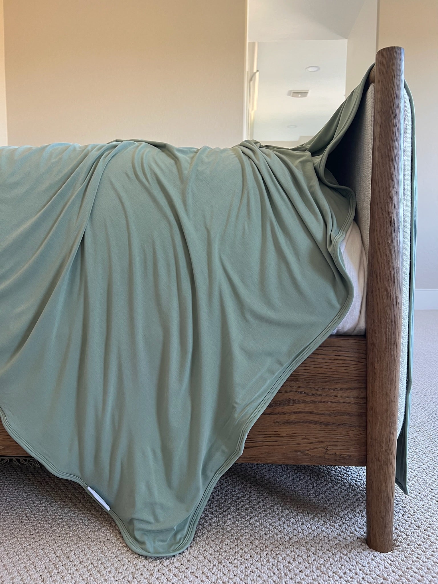 The basil blanket in a rich green hue is casually draped over a wooden bedframe in a bright, carpeted bedroom with neutral walls.