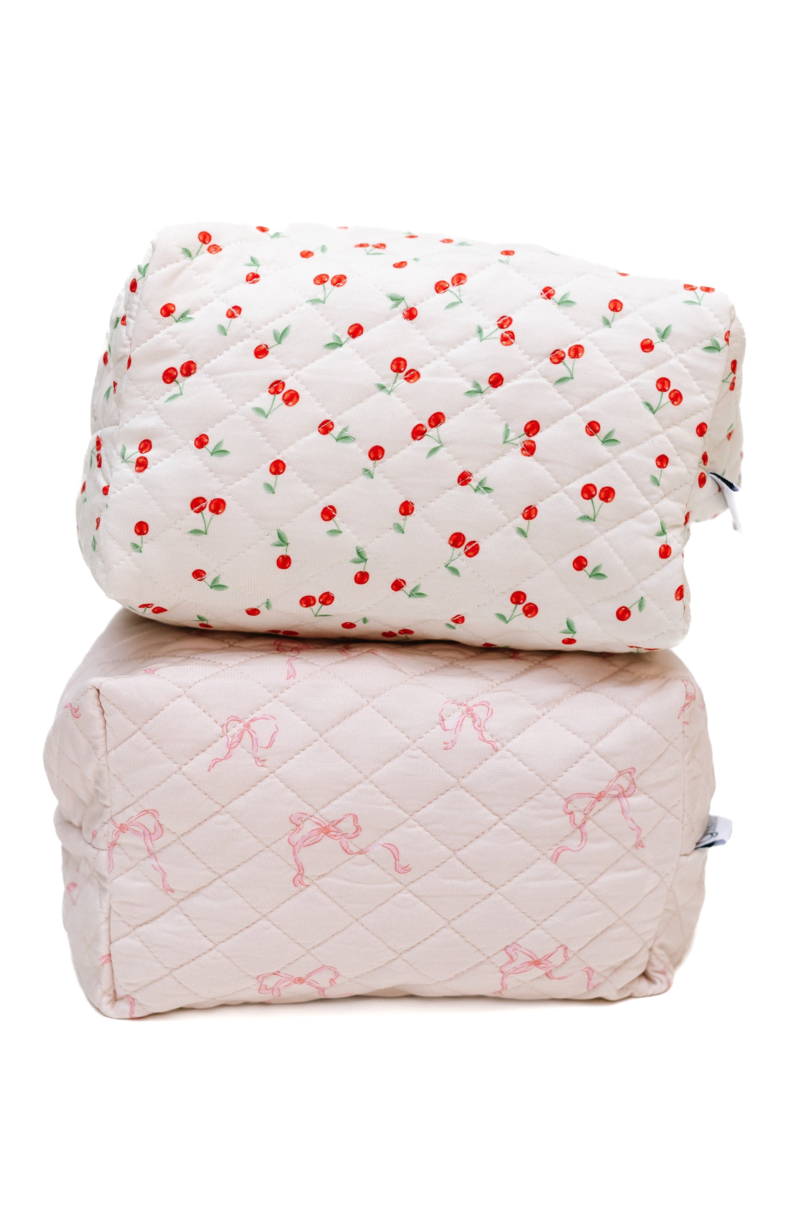 Two quilted, rectangular accessories from the ballerina / travel bag set are stacked: the top is white with a red cherry pattern, the bottom is light pink with bows. Both feature zippers and a diamond stitch texture.