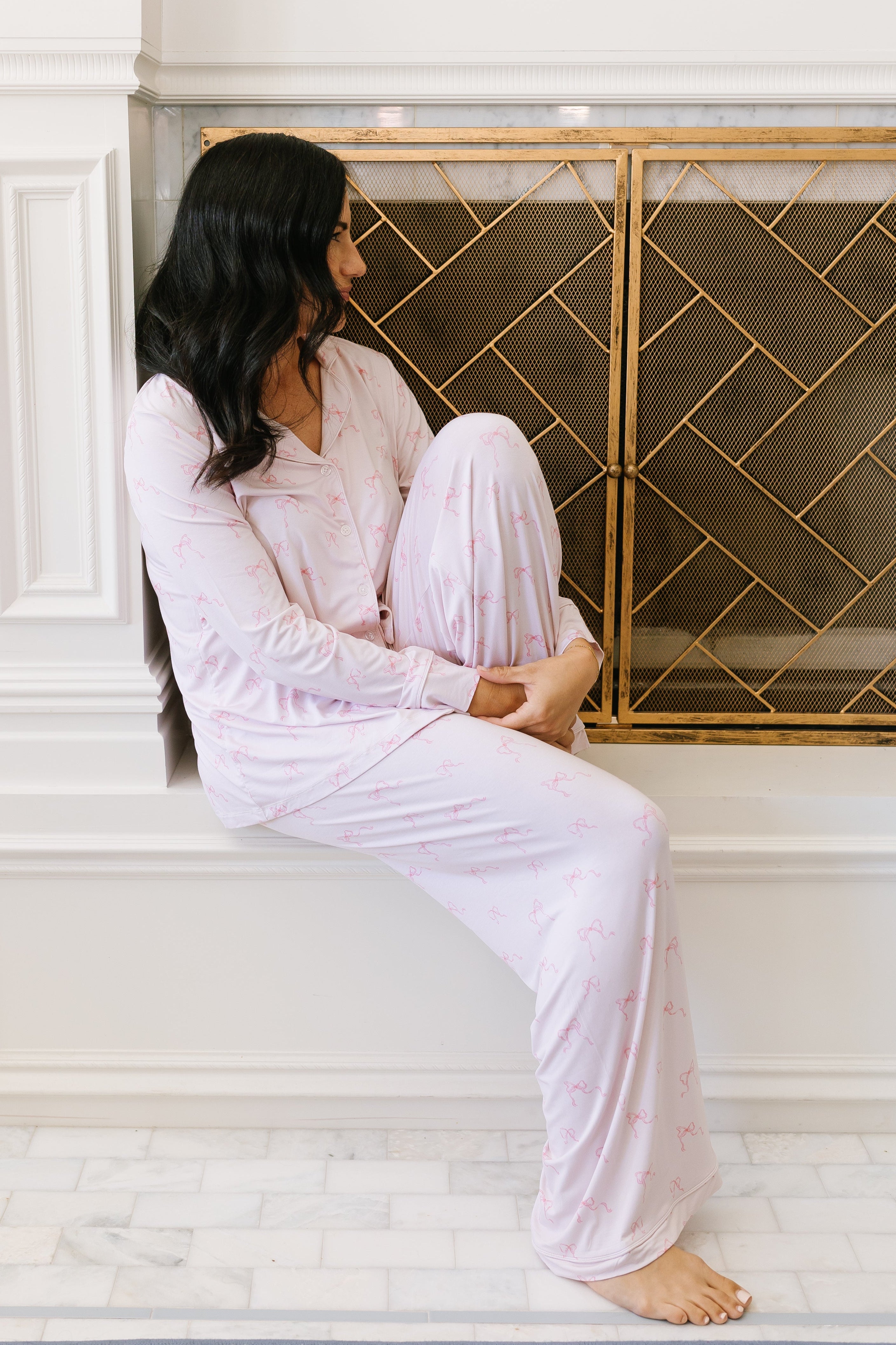 A woman with long dark hair wears the ballerina pajama set / long sleeve with a subtle pattern, sitting on a white fireplace ledge near a gold mesh screen, hugging her knees and gazing to the side.