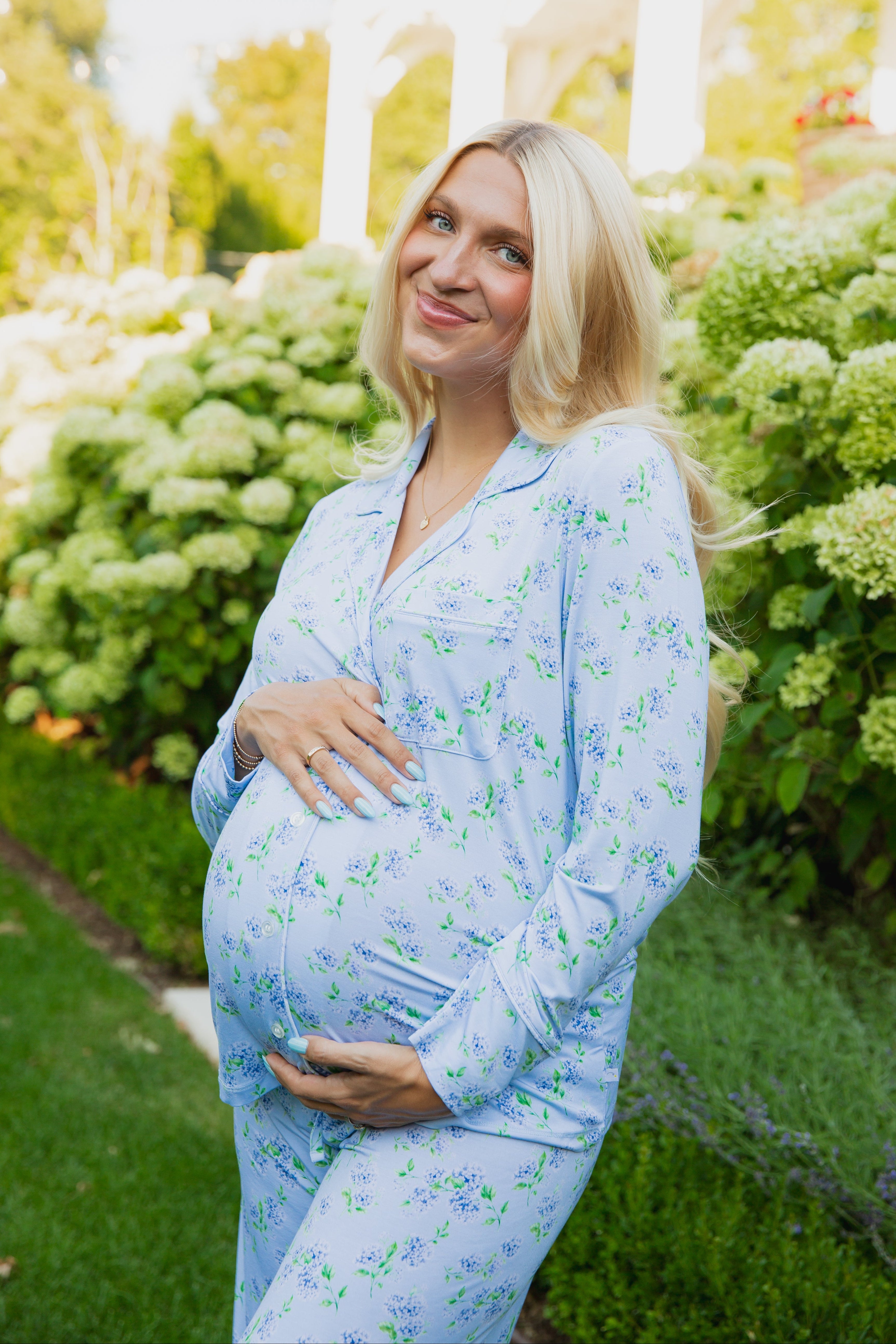 A smiling pregnant woman stands outdoors among lush green bushes and flowers, cradling her belly with both hands. She is wearing the hydrangea long pant pajama set from Sarah Sweeten × Goosebumps.