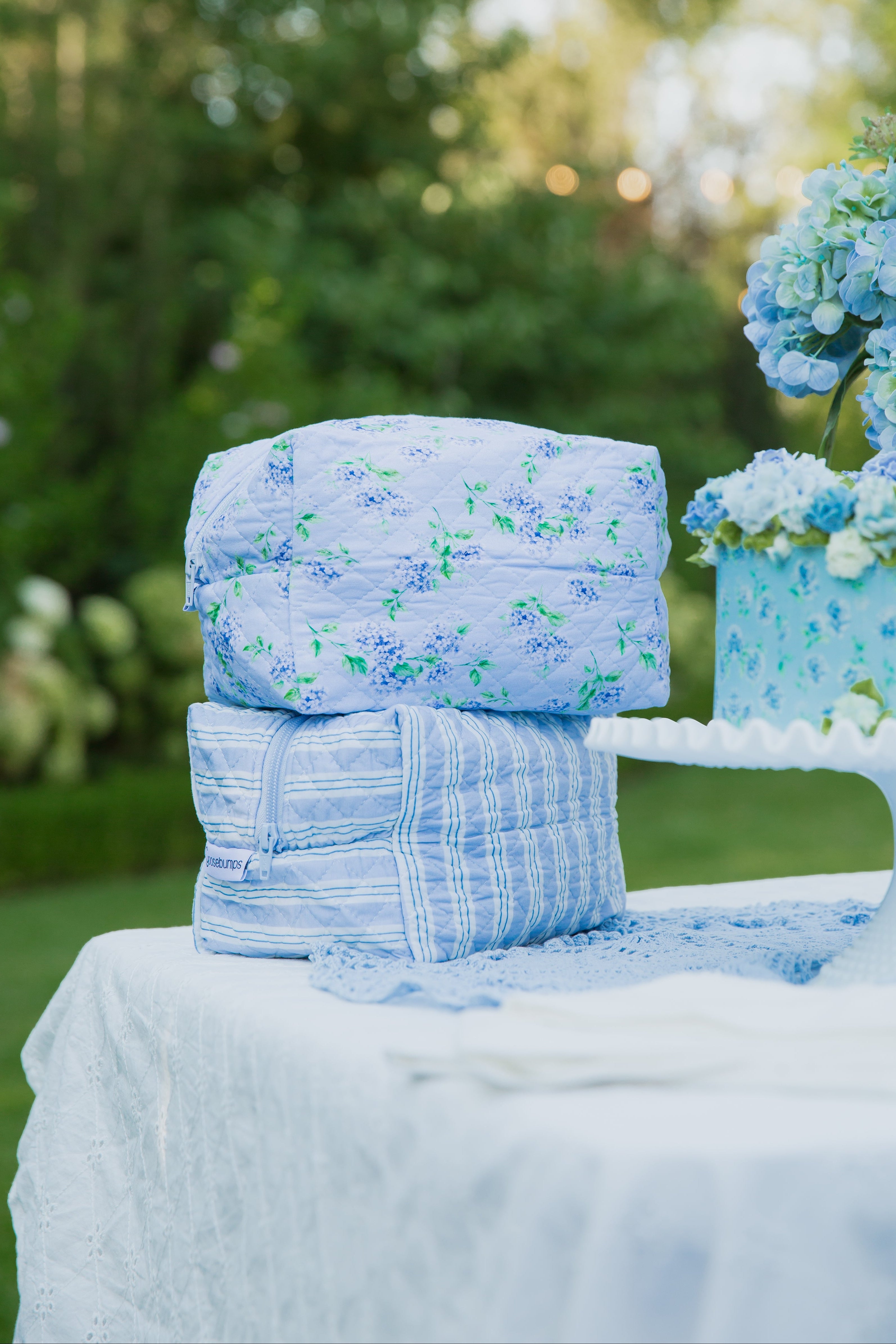 Two blue “hydrangea / travel bag” quilted fabric-wrapped boxes from the Sarah Sweeten Collection sit on a white table outdoors, next to a blue floral cake on a white stand, with greenery and hydrangea print flowers in the background.