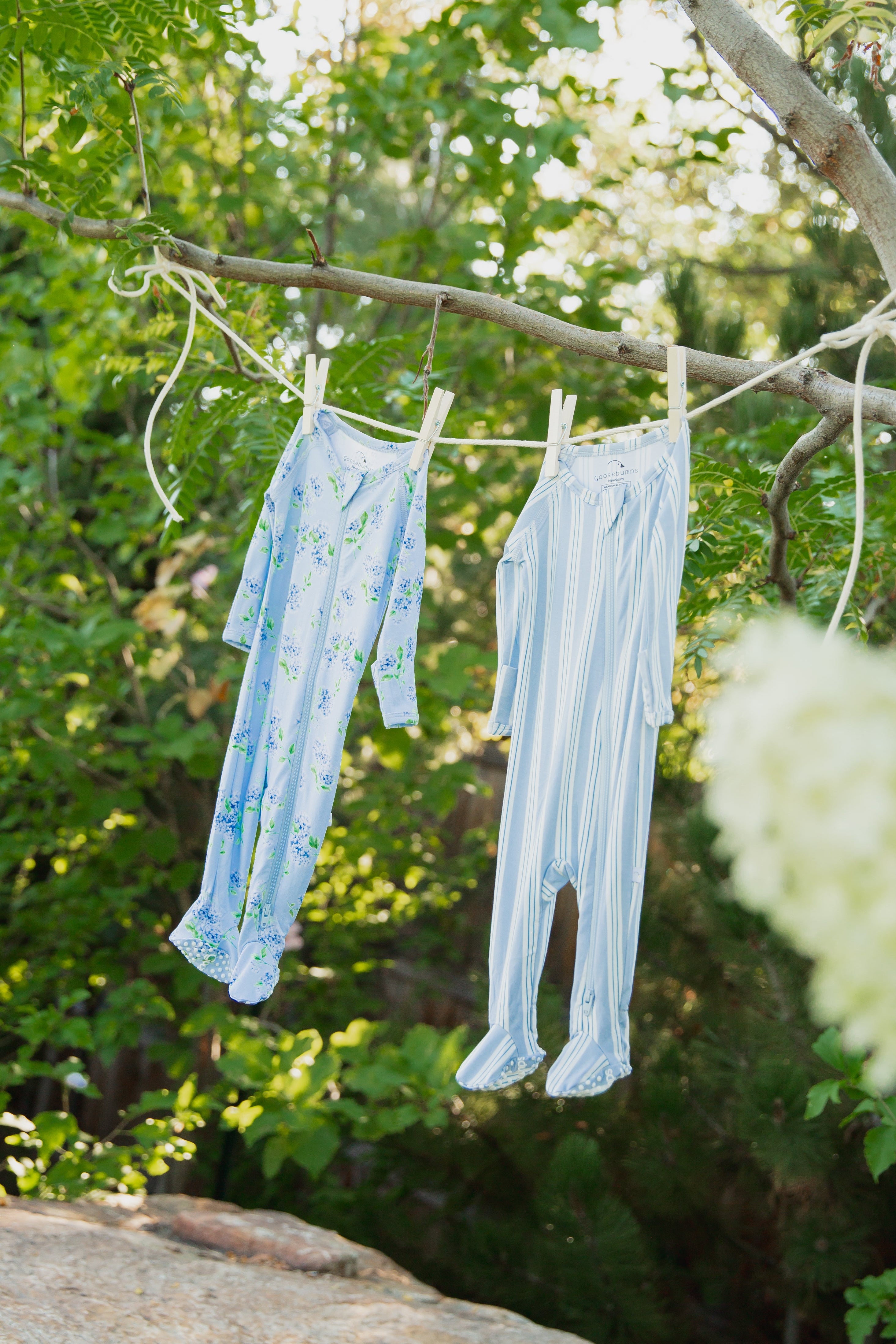Two haven / one piece zipper baby onesies, one with a floral design and one striped, hang outdoors on a clothesline amid green trees and dappled sunlight. Crafted from buttery-soft fabric for all-day comfort, they’re perfect for your little one’s delicate skin.